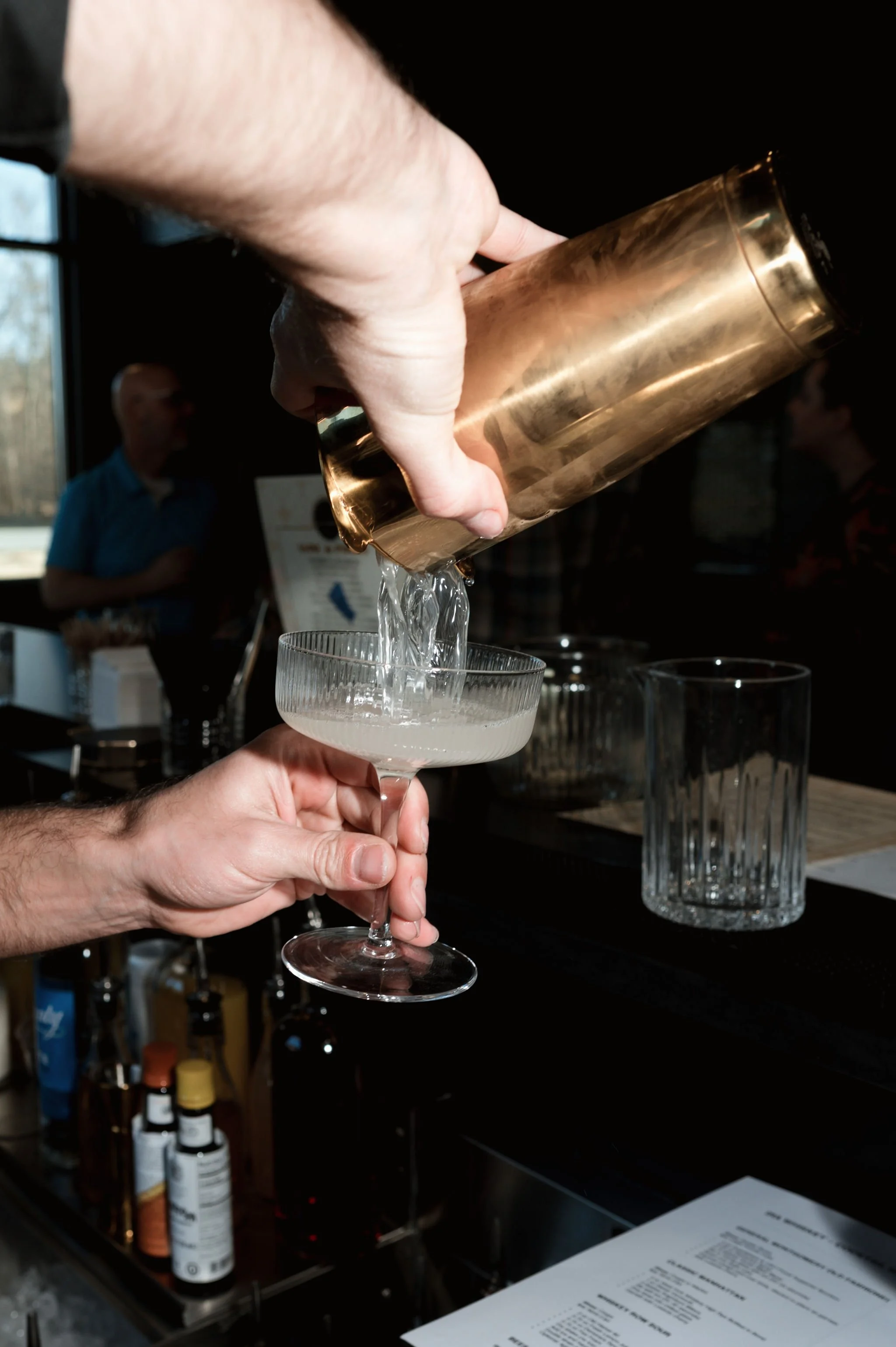 Person pouring a clear liquid from a copper cocktail shaker into a coupe glass