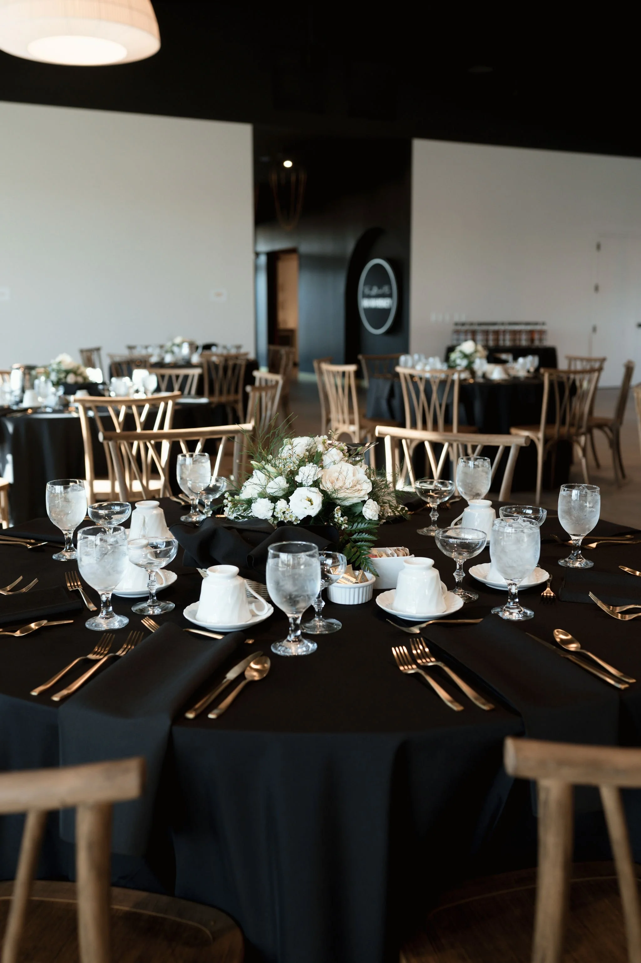 Wedding reception table setup with black tablecloths, floral centerpieces, and formal place settings in a decorated banquet hall.