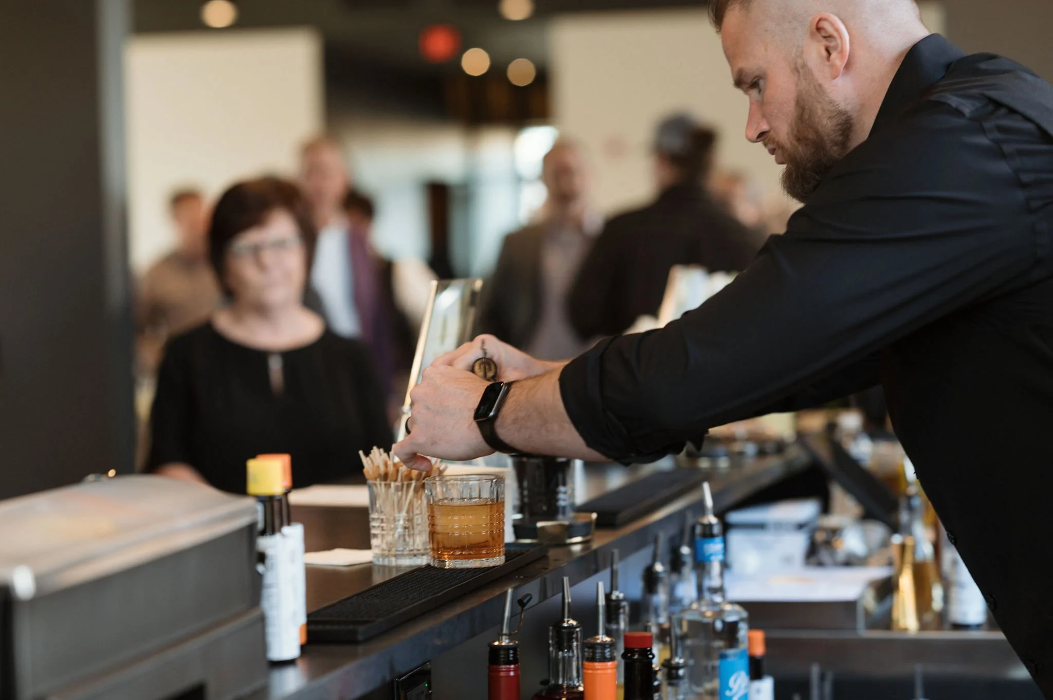 A man with a beard in a black shirt is pouring a beverage into a glass at a counter during a social event or gathering, with blurred people in the background.