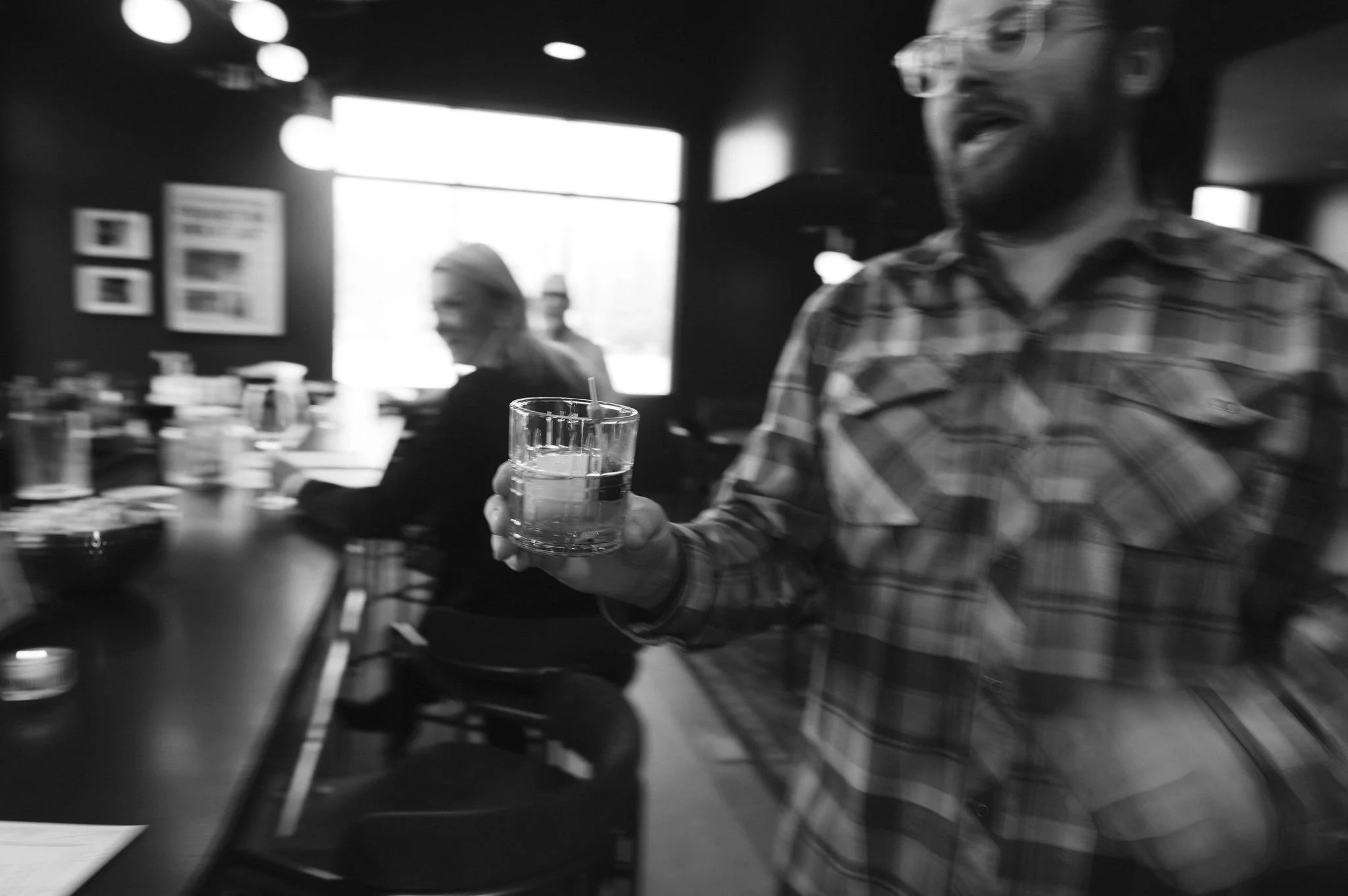 A black and white photo capturing a man in a plaid shirt sitting at a table in a dimly lit restaurant or cafe, holding a glass of water with a straw. In the background, a woman is visible, and the scene includes decorative light fixtures and framed p