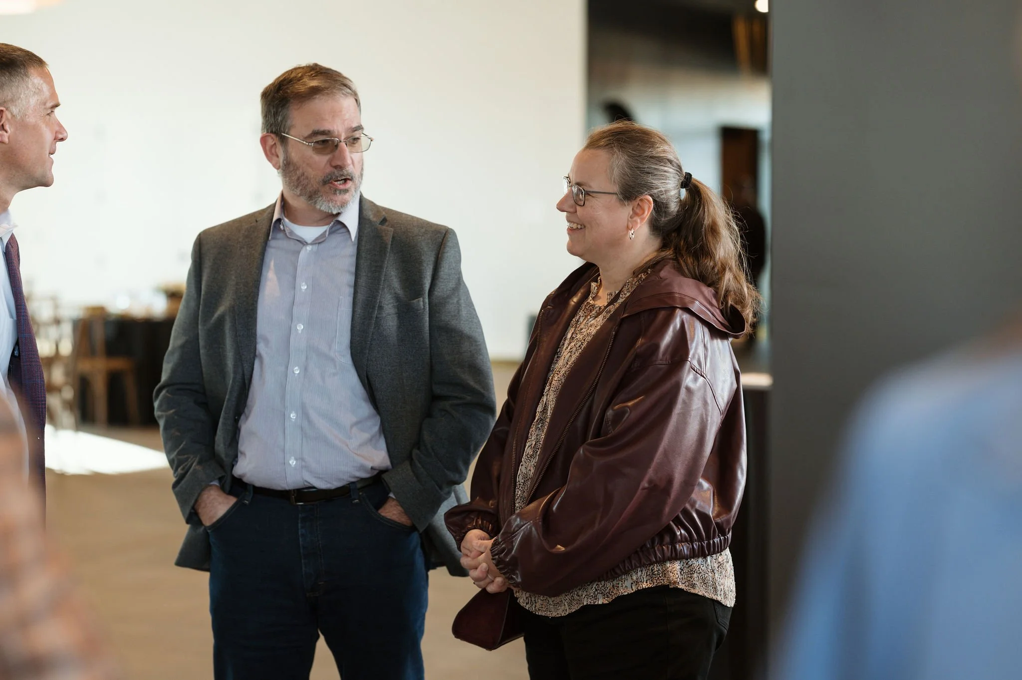 Three people engaged in conversation indoors, with two men and one woman, all dressed casually and smiling.