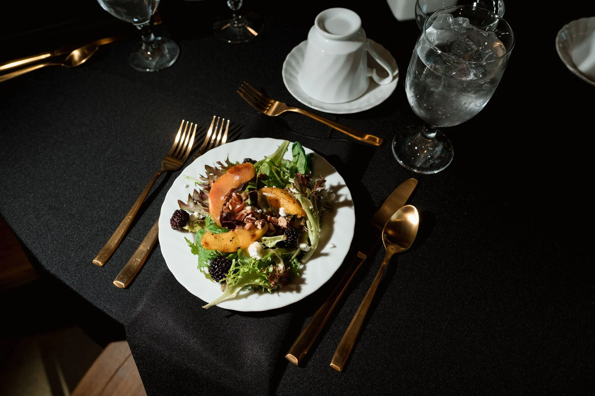 A black tablecloth with a white plate of salad containing greens, blackberries, and sliced peaches, gold utensils, a glass of water with ice, two wine glasses, and a white coffee cup and saucer.