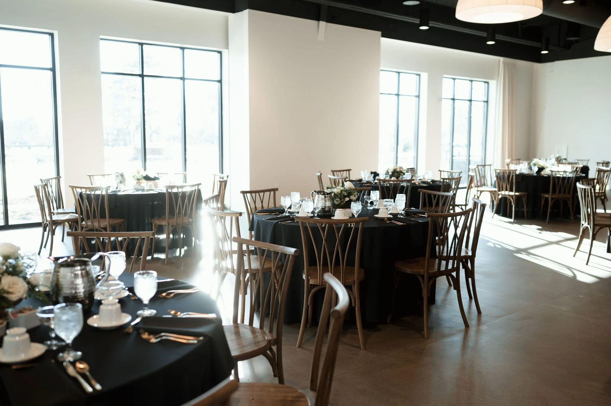 An elegantly set dining area with round tables covered in black tablecloths, adorned with floral centerpieces and glassware, surrounded by wooden chairs with curved backs, in a bright room with large windows and modern lighting fixtures.