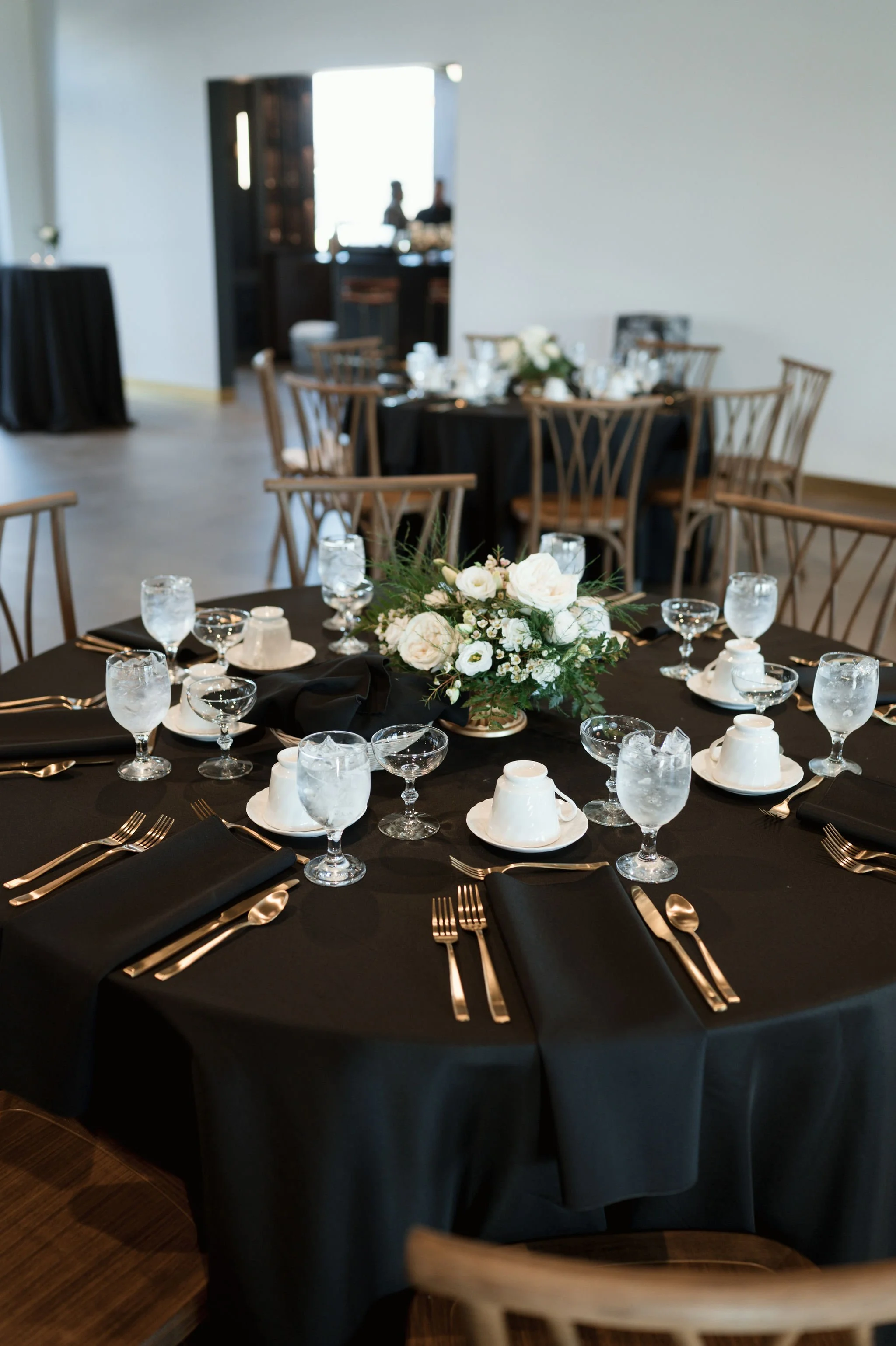 Elegant round dining table set for a formal event with black tablecloth, white floral centerpiece, black napkins, clear water glasses, white cups, and gold flatware, in a bright room with wooden chairs.