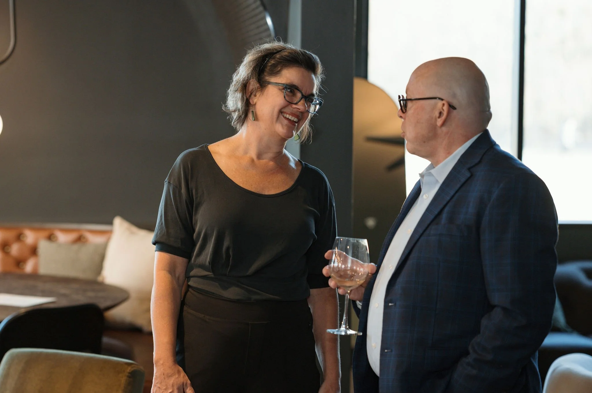 A woman with short gray hair and glasses smiling and holding a wine glass, talking to a bald man with glasses and a suit in an indoor setting.