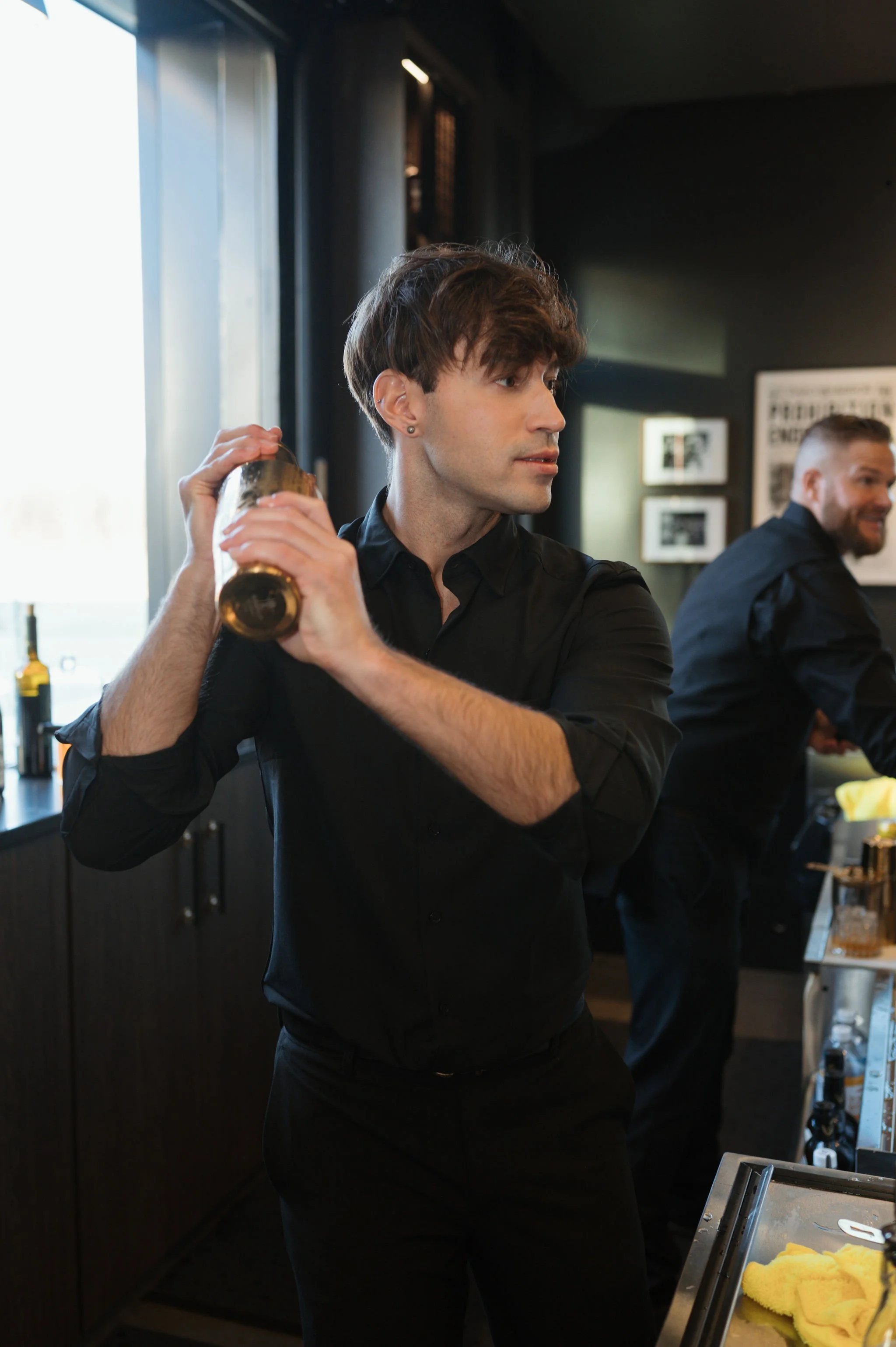 A young man with dark hair, dressed in a black shirt, holding a cocktail shaker, appears to be preparing a drink behind a bar in a dimly lit setting with another man in the background.