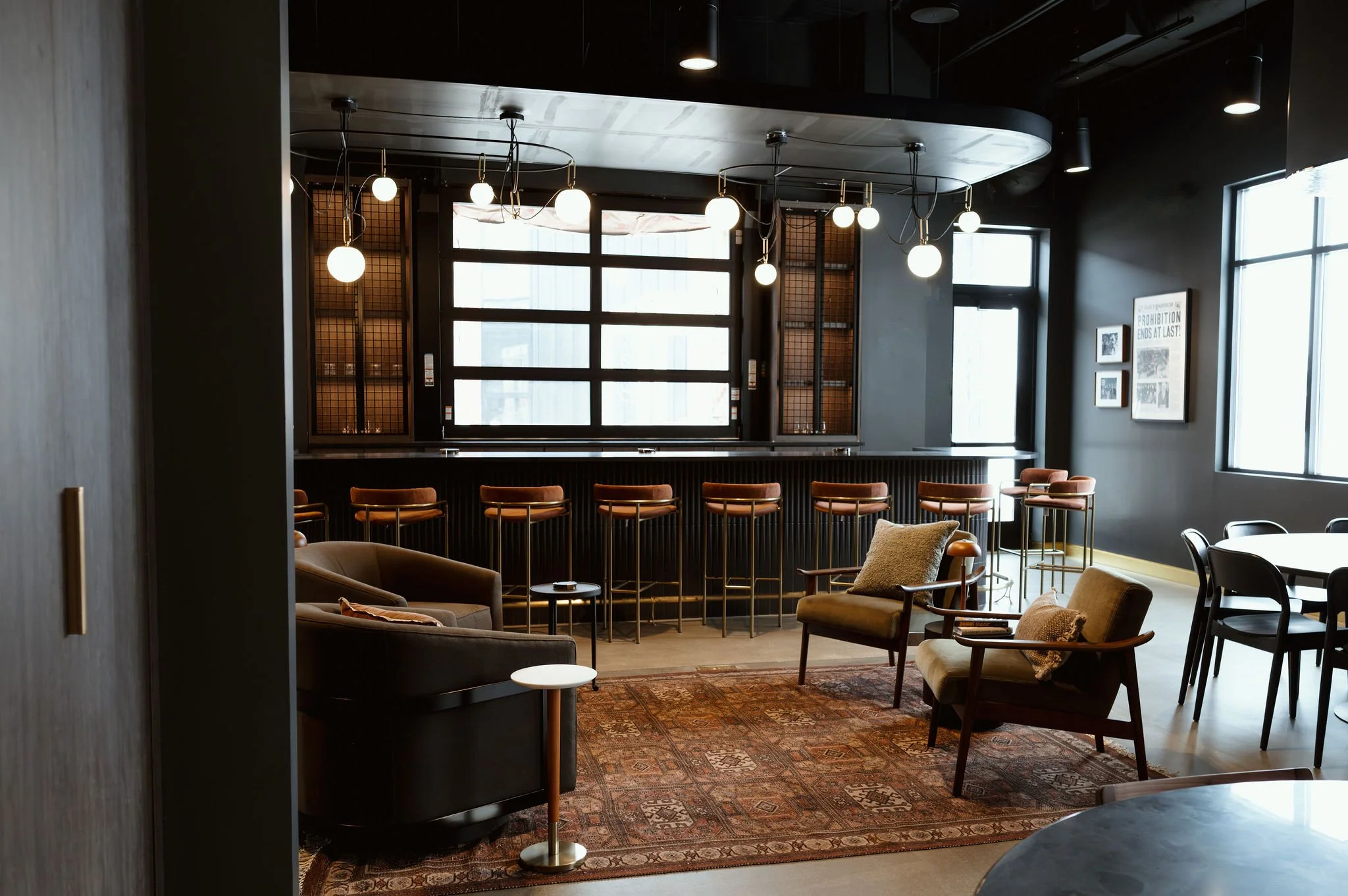 Interior of a modern, stylish lounge or cafe with large windows, hanging globe and cylindrical lights, armchairs, sofas, and patterned area rugs.