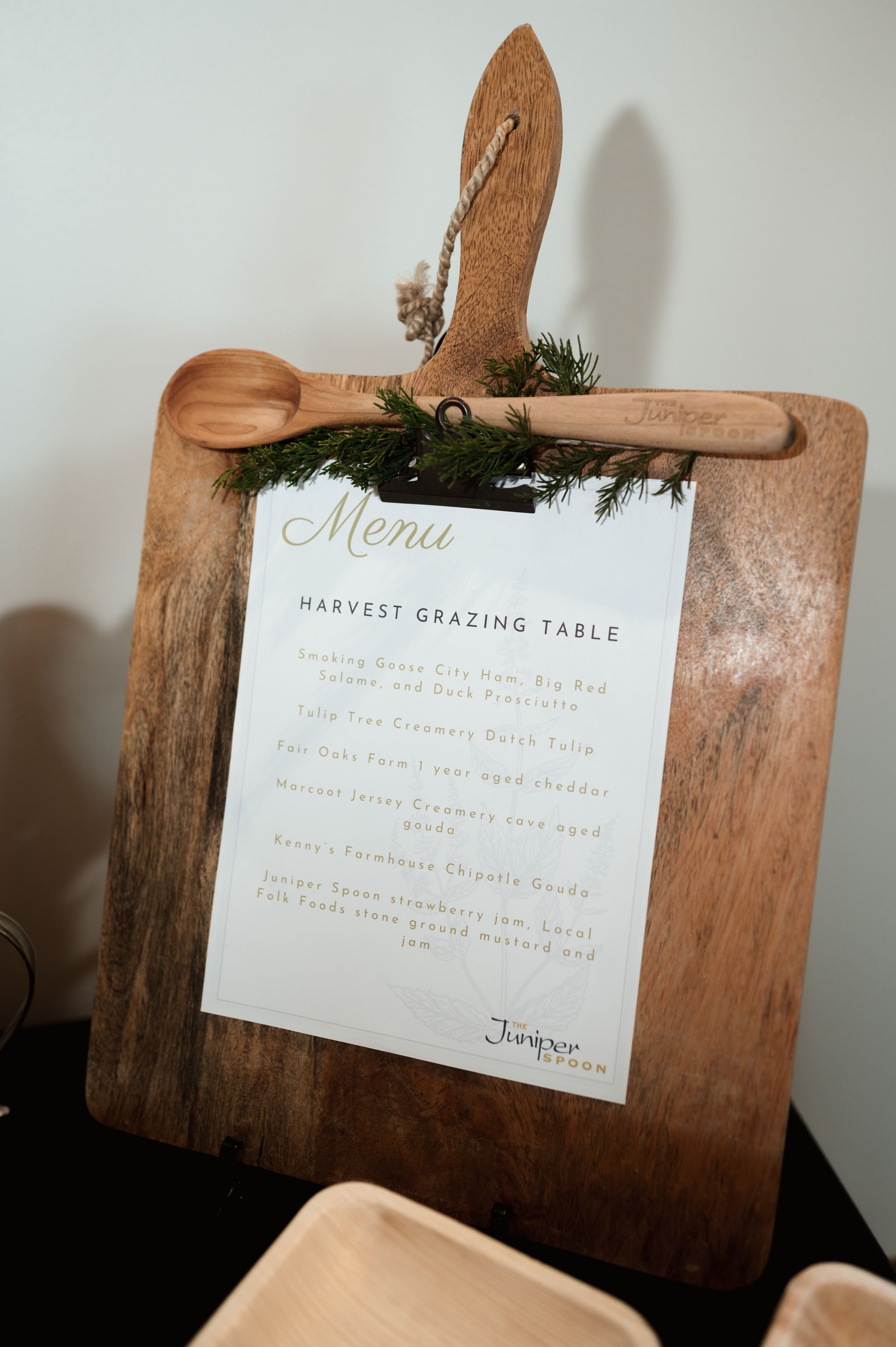 A wooden signboard displaying a menu titled 'Harvest Grazing Table' with food items listed below, adorned with small evergreen sprigs, a wooden spoon and a wooden high heel shoe decorative piece attached at the top.