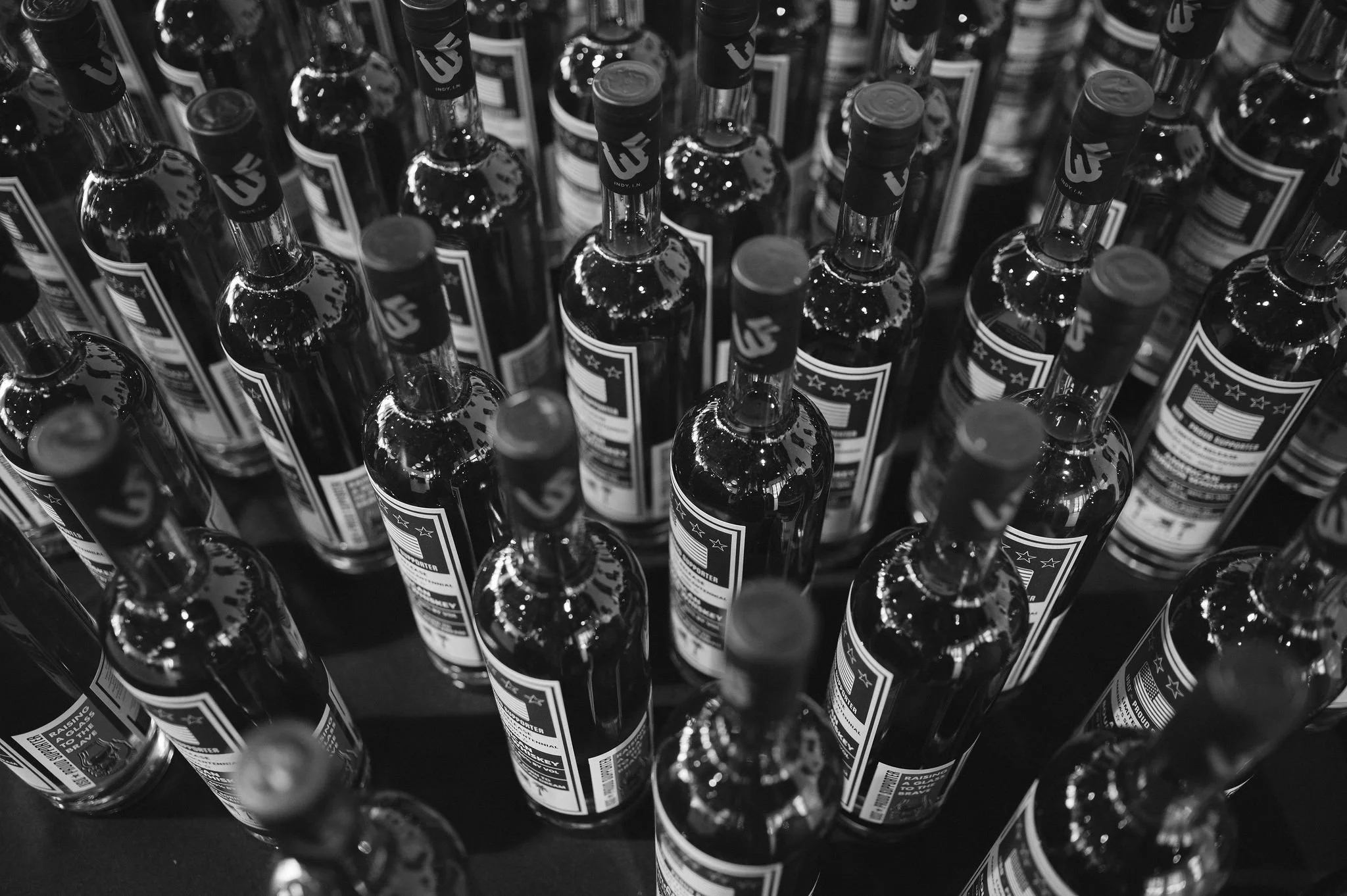 Black and white photo of multiple bottles of alcohol arranged in rows with labels visible.