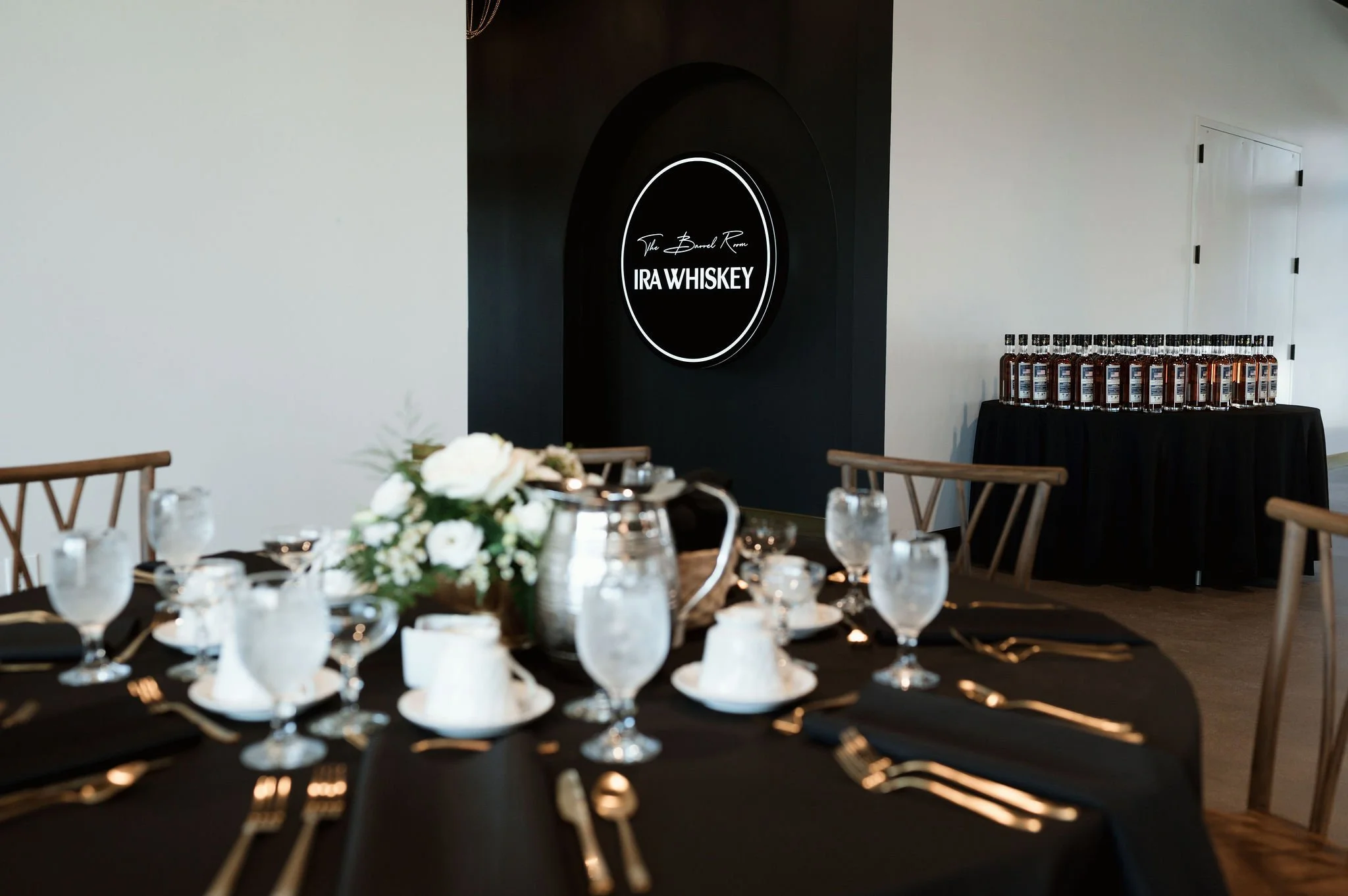 A banquet table set with glassware, plates, and a floral centerpiece, in front of a black wall with a neon sign for IRA Whiskey, and a table with bottles of whiskey in the background.