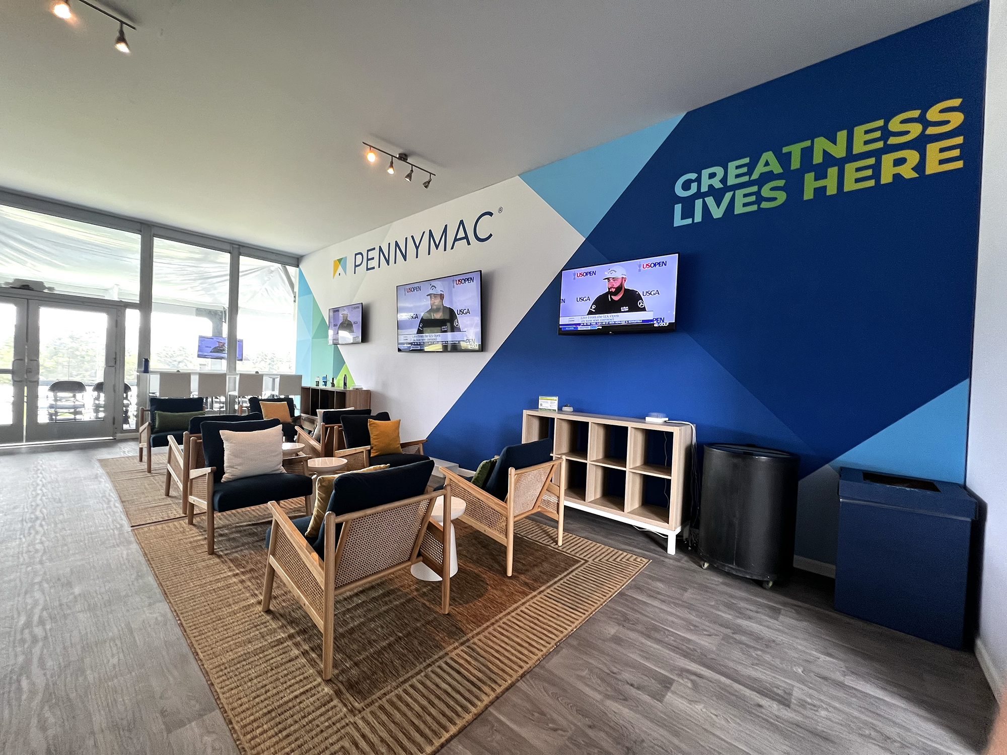 Indoor seating area with chairs, a rug, and a wall with 'PENNYMAC' sign and 'GREATNESS LIVES HERE' message, TV screens displaying sports and news, near large windows.