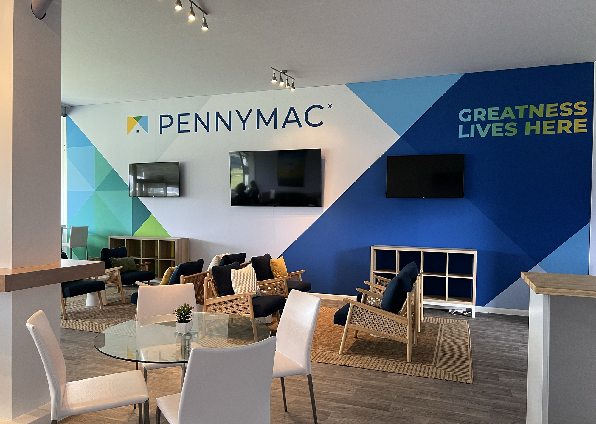 Interior of a modern office lounge or waiting area with colorful geometric wall art, flat-screen monitors, chairs, tables, and a small plant.