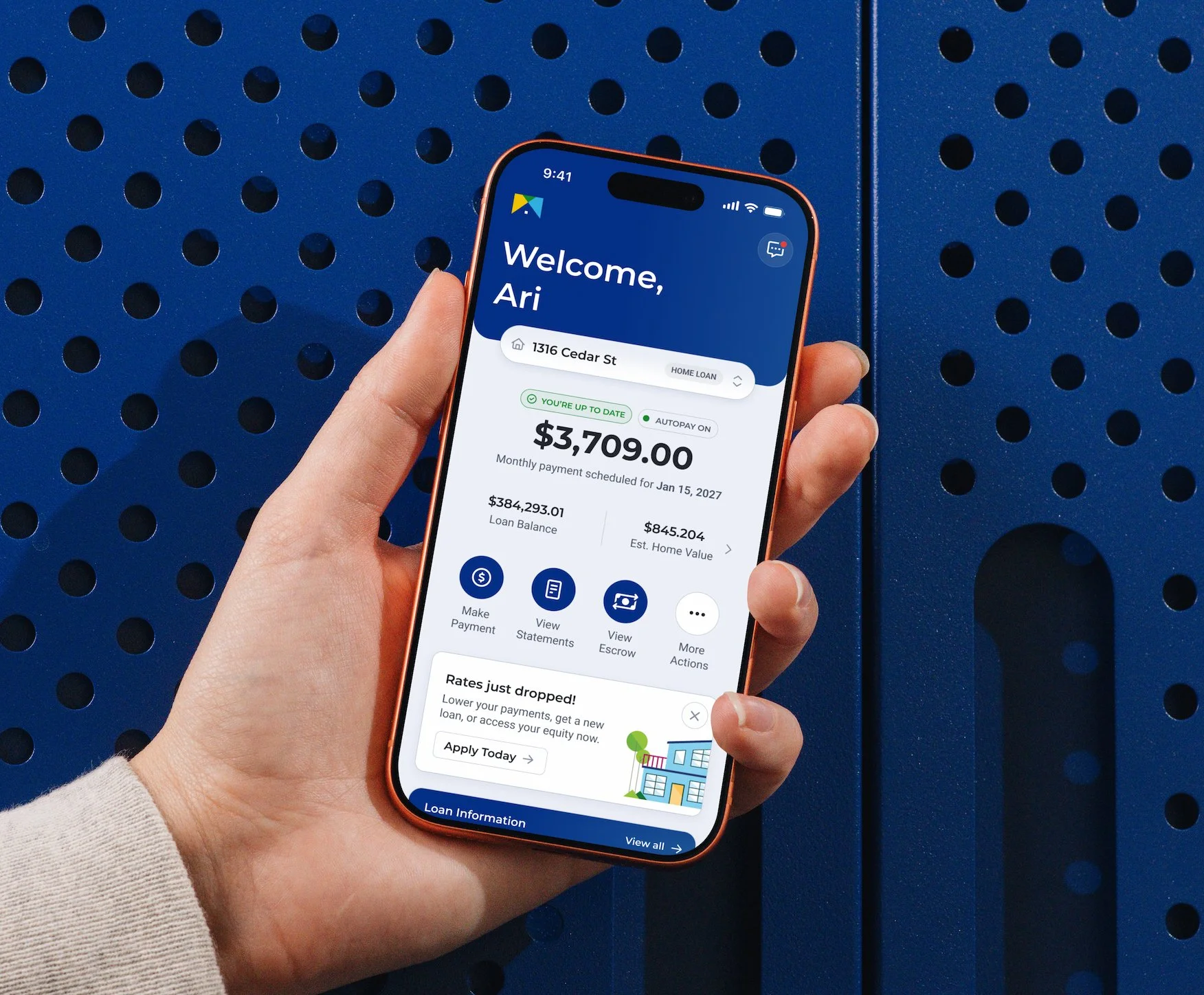 A person's hand holding a smartphone displaying a home loan management app with details including loan balance, home value, monthly payment, and options to view statements and make payments, against a blue perforated surface background.