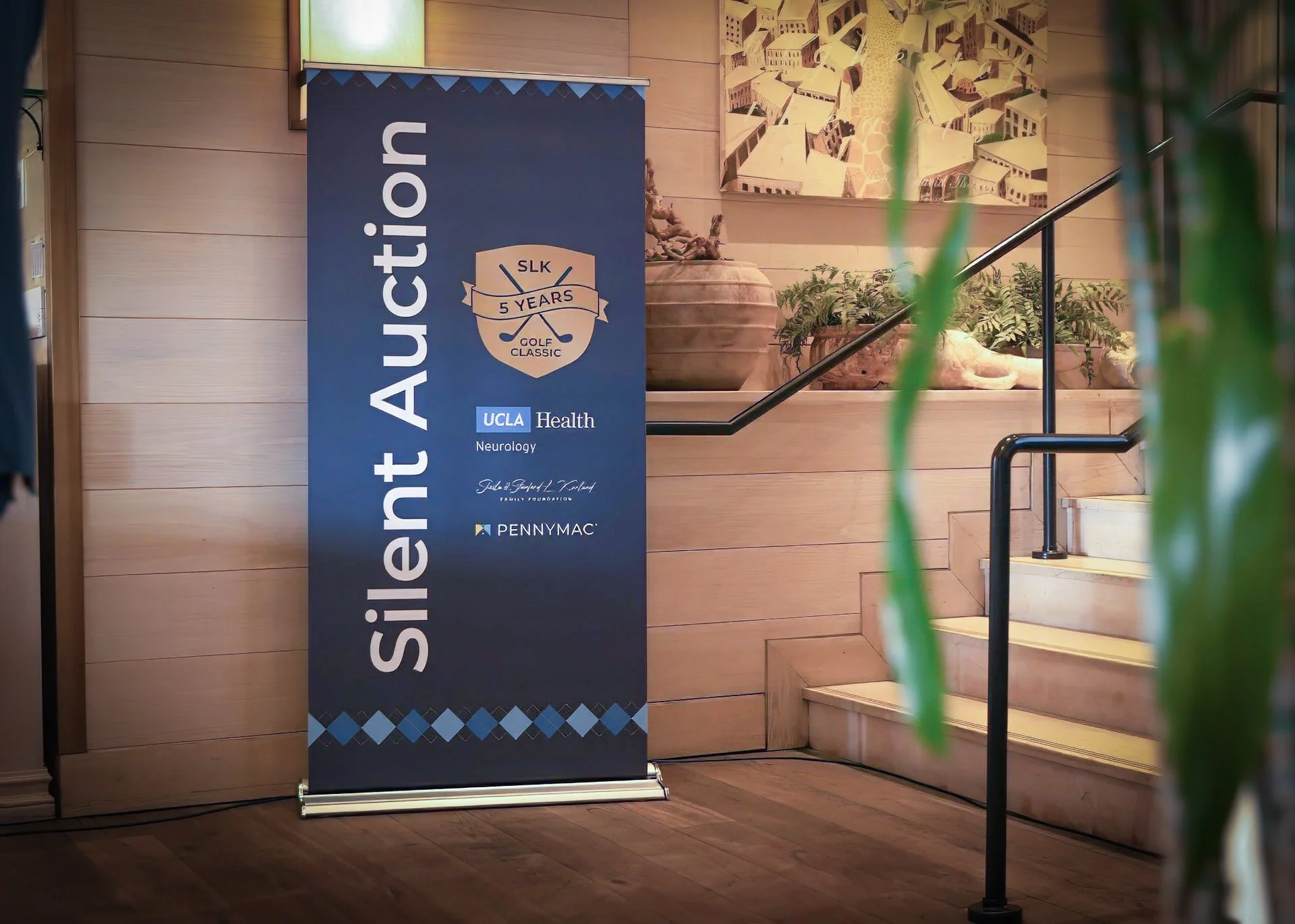 A promotional banner on a floor stand for the Silicon Autction, celebrating 5 years of SLK Golf Classic, affiliated with UCLA Health Neurology, displayed on a wooden wall near a staircase with plants nearby.