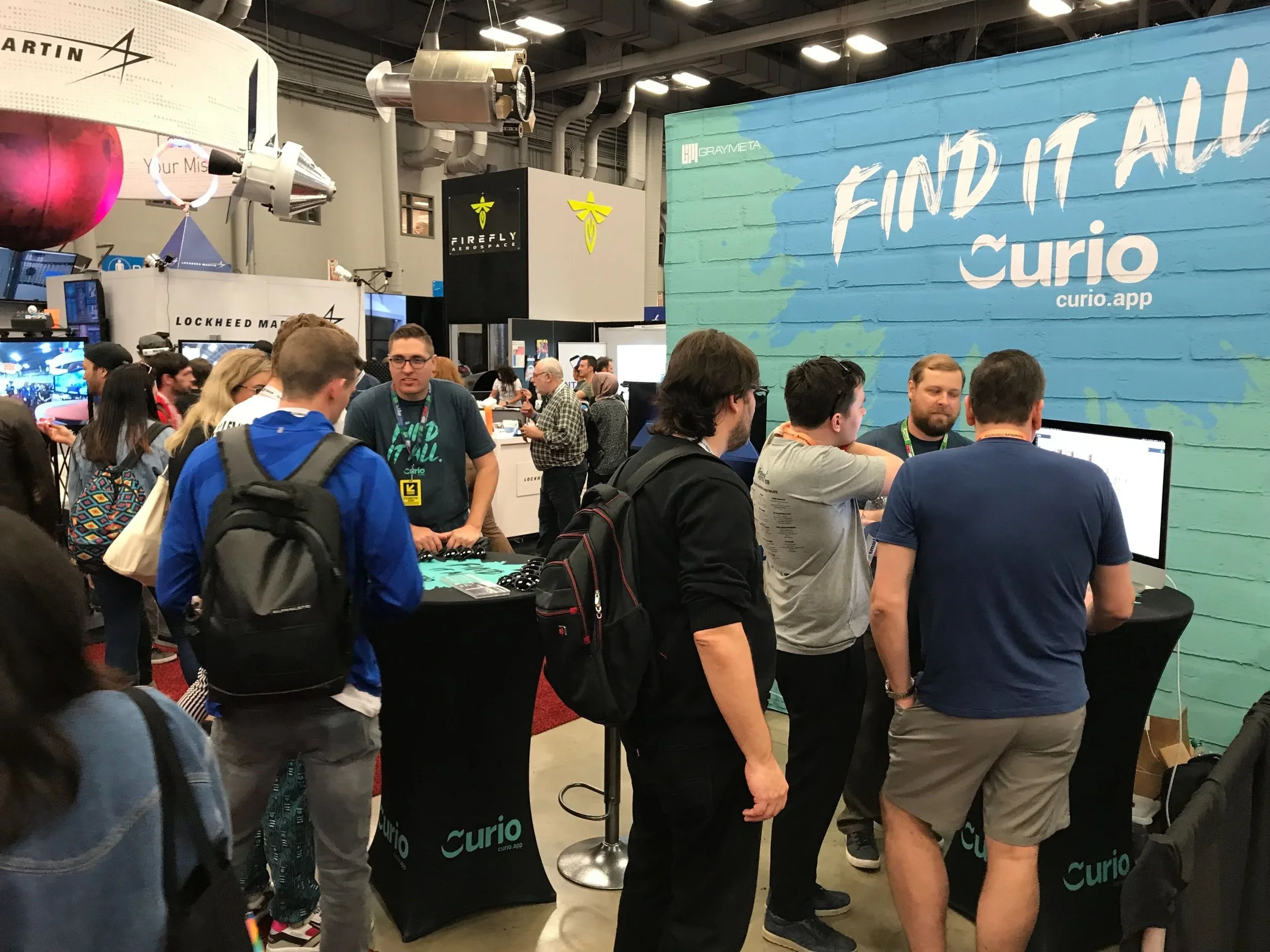 Crowd of people at a convention or expo, gathered around a booth with a large blue and green sign that says "FIND IT ALL" and "curio.app." Several individuals are engaging with displays and technology, with some wearing lanyards and backpacks.
