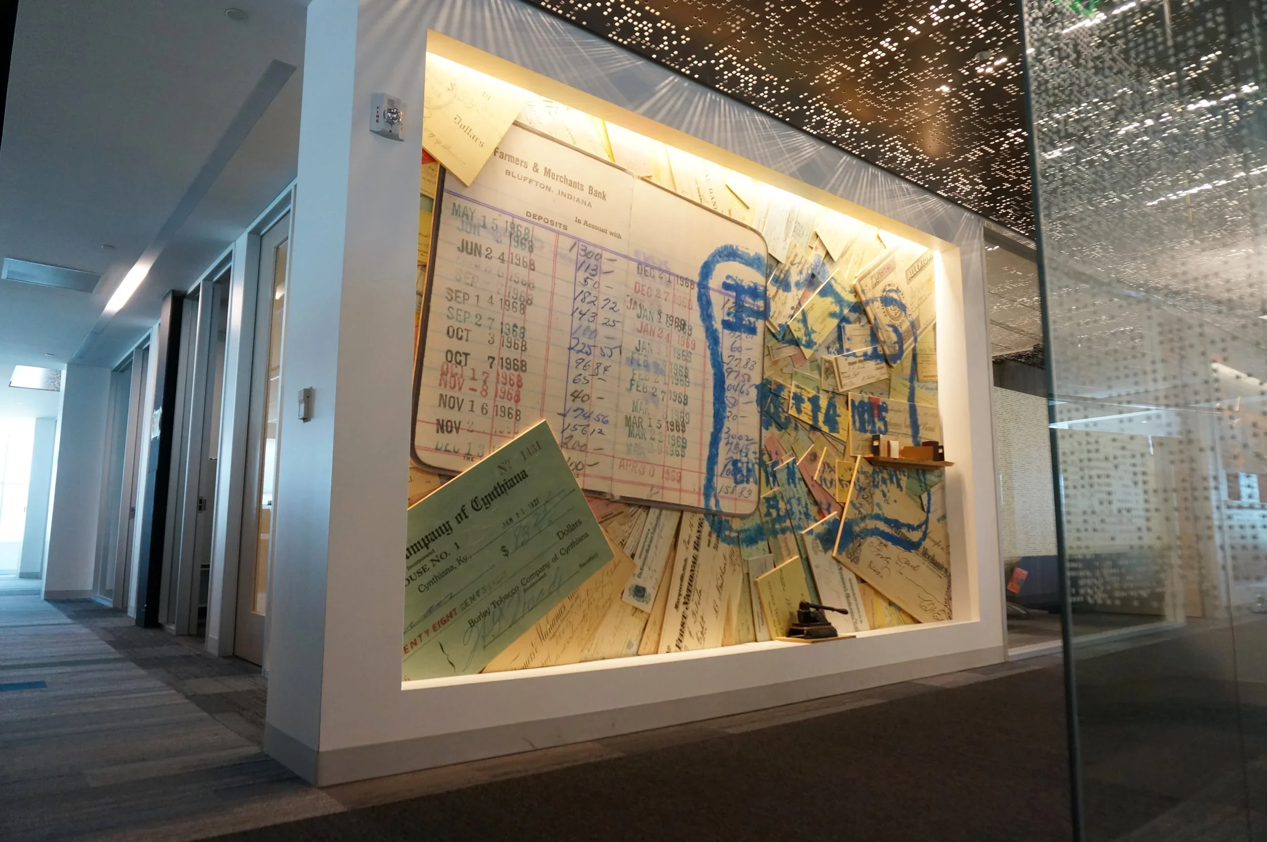 A display case with historical documents, including old bank deposit slips and handwritten notes, arranged on a wall with a modern ceiling and windowed walls in the background.
