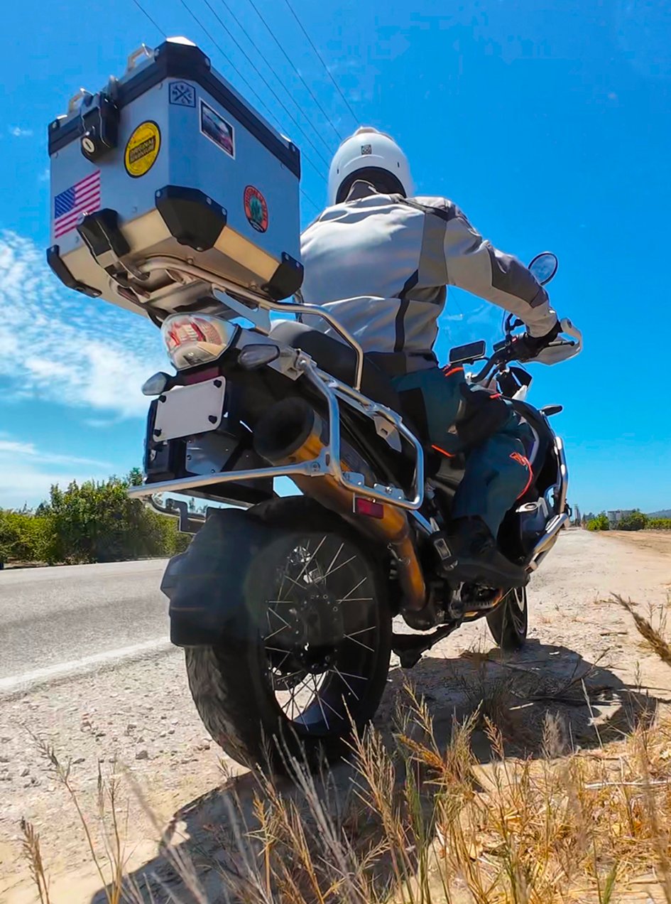 A person riding a motorcycle on a dirt road on a sunny day, wearing a white helmet and a white jacket, with a large box attached to the back of the motorcycle decorated with stickers, including American flags.
