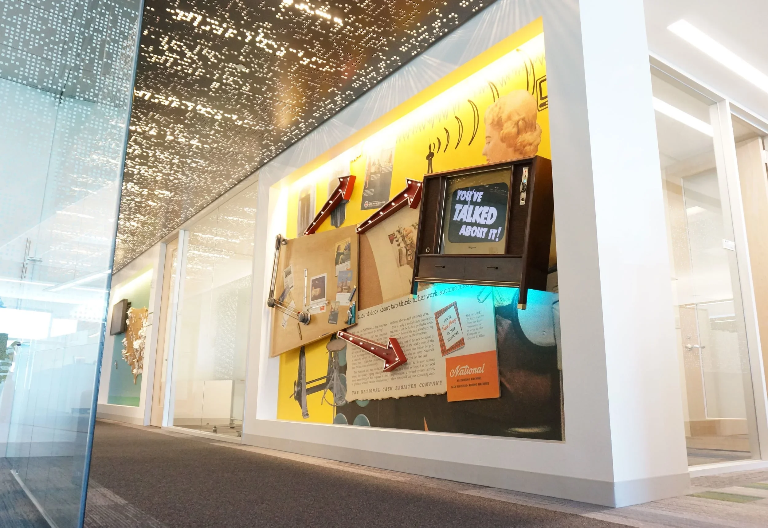 Interior of a modern building with a decorative ceiling, a large yellow board displaying vintage advertisements, an old television with the text 'You've Talked About It!', and informational posters on a corkboard.