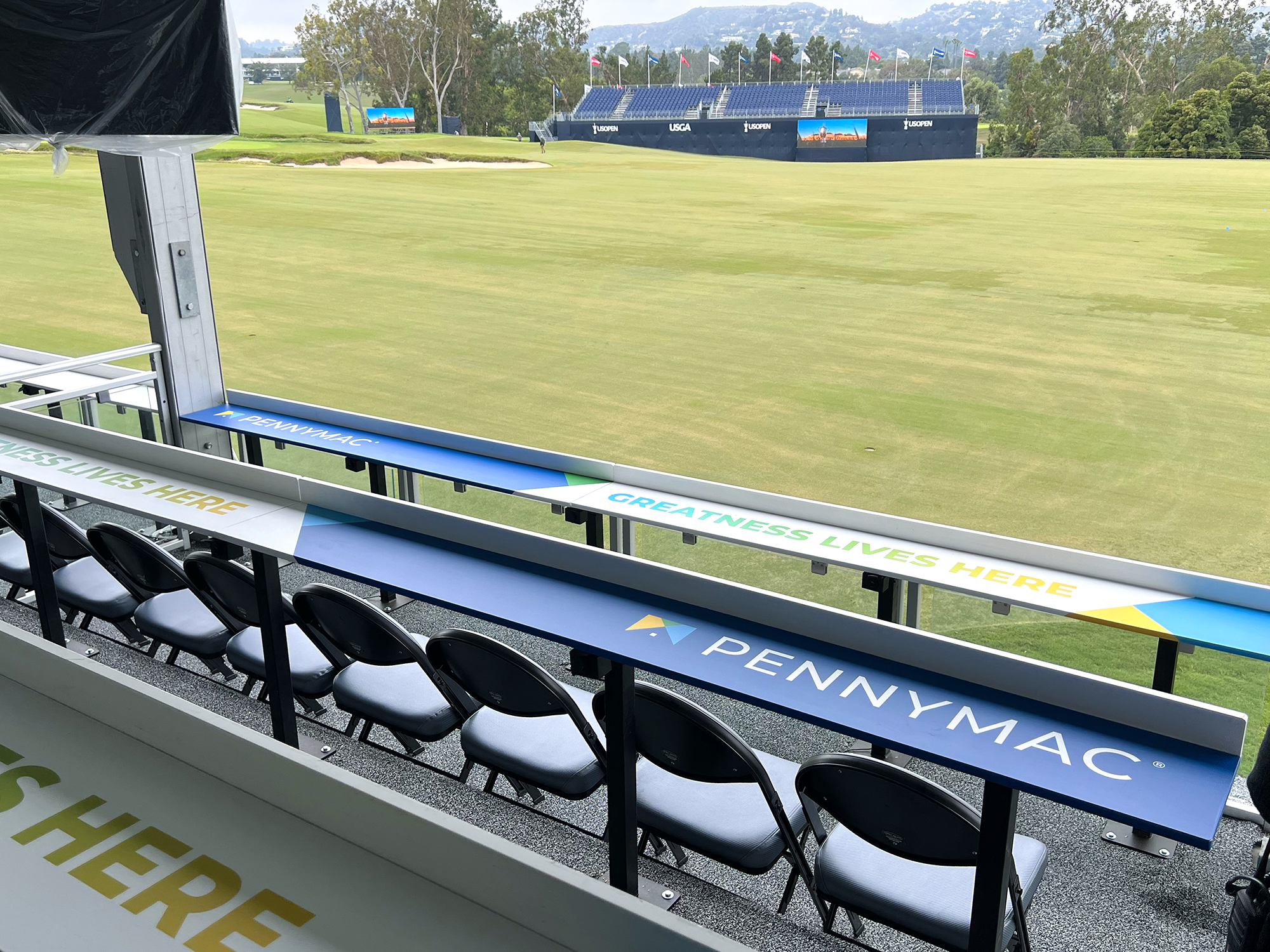 View from the spectator seating area at a golf course with green grass, trees, hills, and blue sky, showing the fairway, sand traps, digital screens, and flags at the US Open golf tournament.