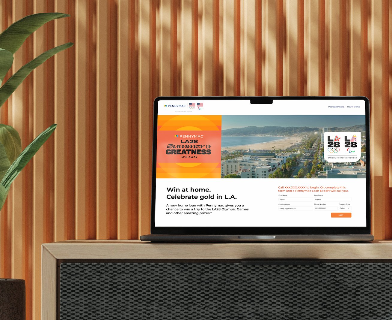 Laptop displaying a promotional webpage for PennyMac's LA28 Summer of Greatness Giveaway, with a scenic photo of Los Angeles and Olympic logos, placed on a wooden table with a plant nearby, against a wooden slat wall.