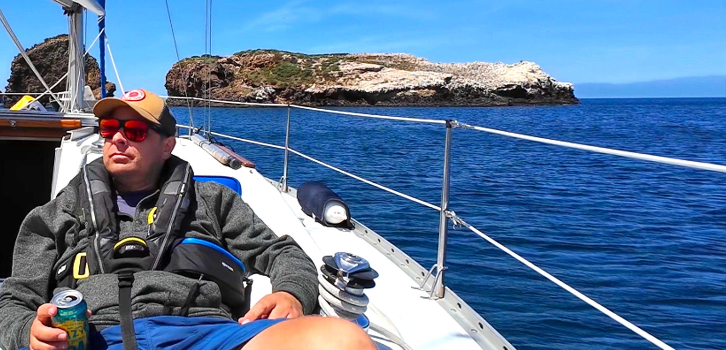 A man wearing sunglasses, a tan cap, and a life jacket relaxing on a sailboat with a rocky island in the background and clear blue sky.
