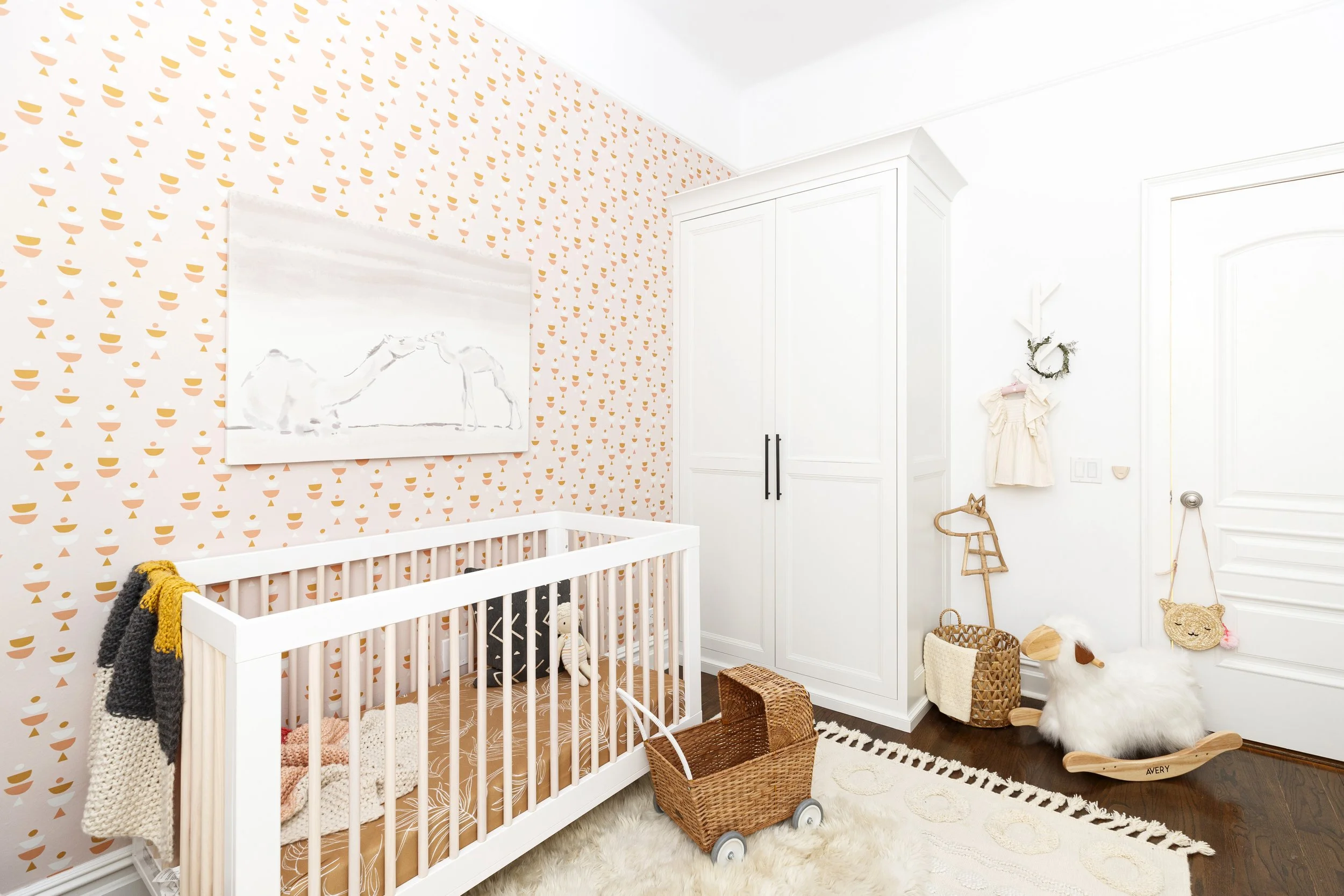 white crib with mustard bedding, rattan accents and white custom closet against pink wallpaper in Manhattan baby girl nursery designed by Westchester, NY interior designer Jordana Ross Interiors