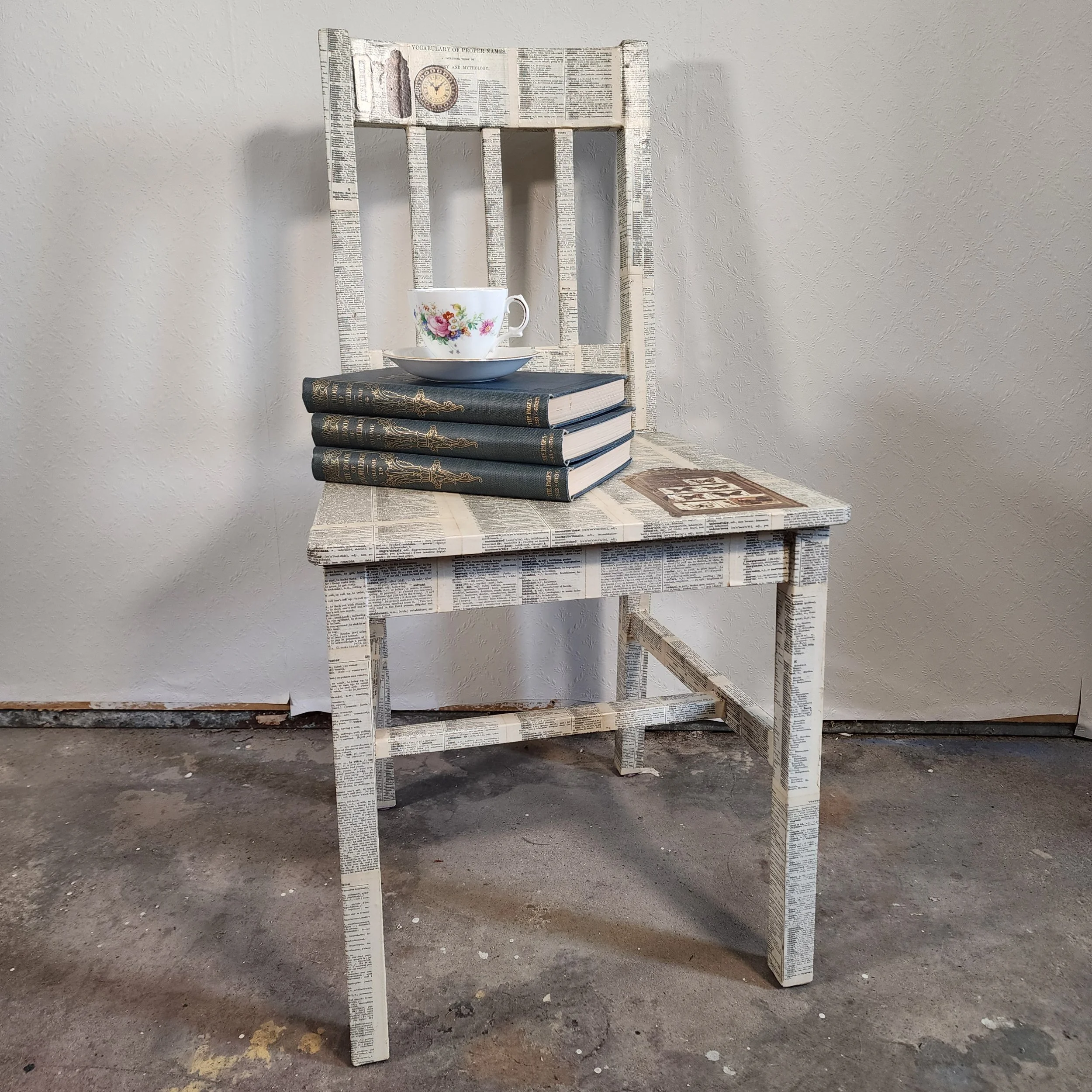 A chair with a newspaper print design, placed against a plain wall, with three books, a decorative bowl with a floral mug on top, and a small decorative item on the seat.
