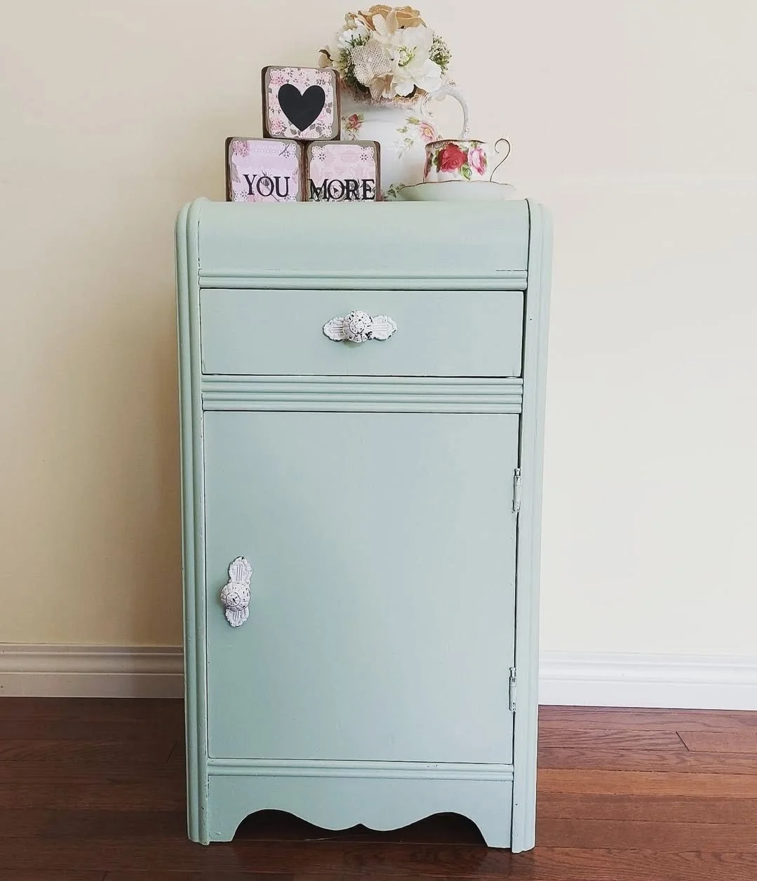 A light blue vintage cabinet with decorative white and pink knobs, topped with a floral arrangement, a pink and white teacup, and framed words saying "You" and "More" with a black heart.