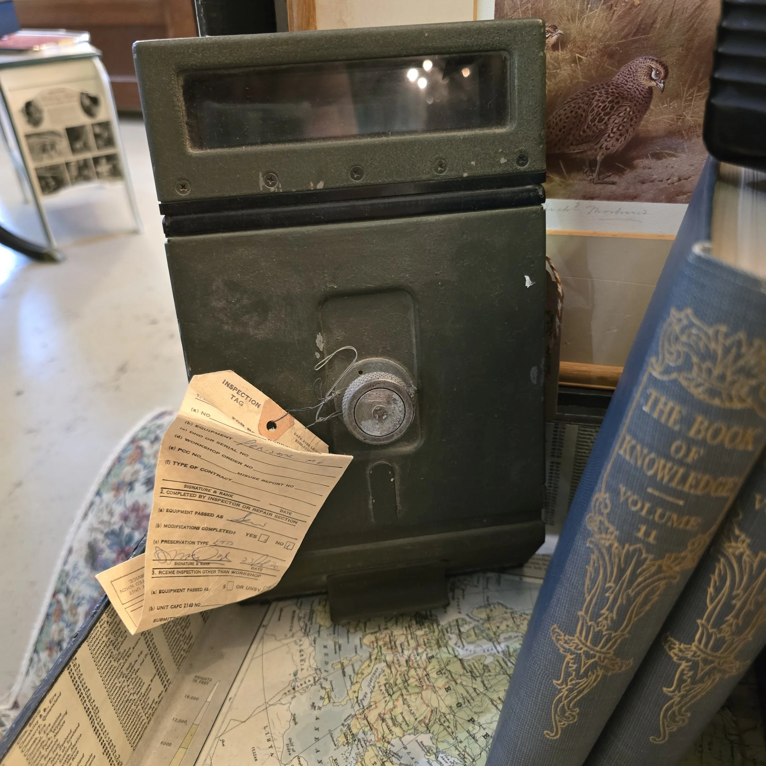 An old black safe with a round lock and a slot on top, placed on a table with maps and books nearby. The table has a map, and there is a book titled "The Book of Knowledge, Volume 11" beside the safe. In the background, there is a poster of a bird an