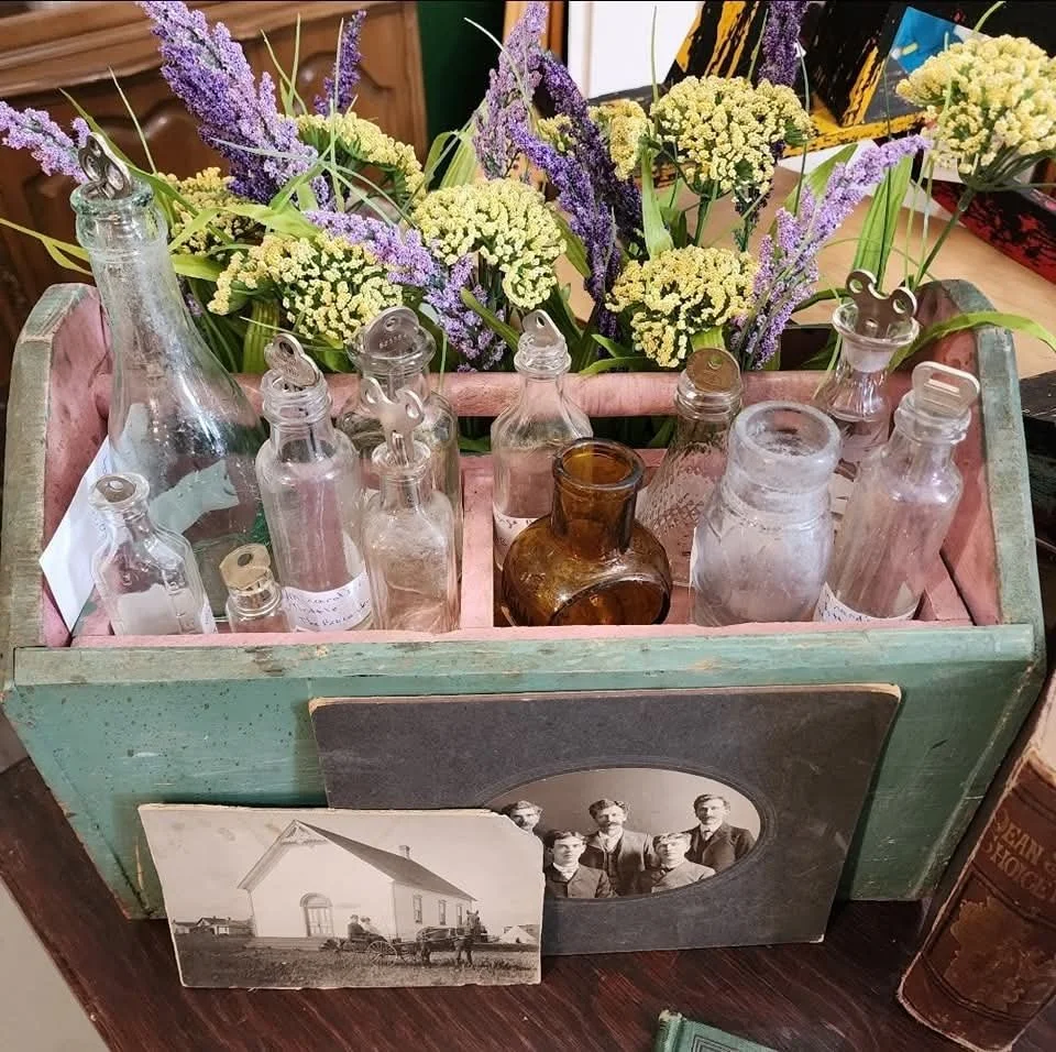 A vintage display with empty glass bottles in a wooden tray, fresh purple and green flowers behind, and old black-and-white photographs of a house and a group of people, all arranged on a wooden surface.