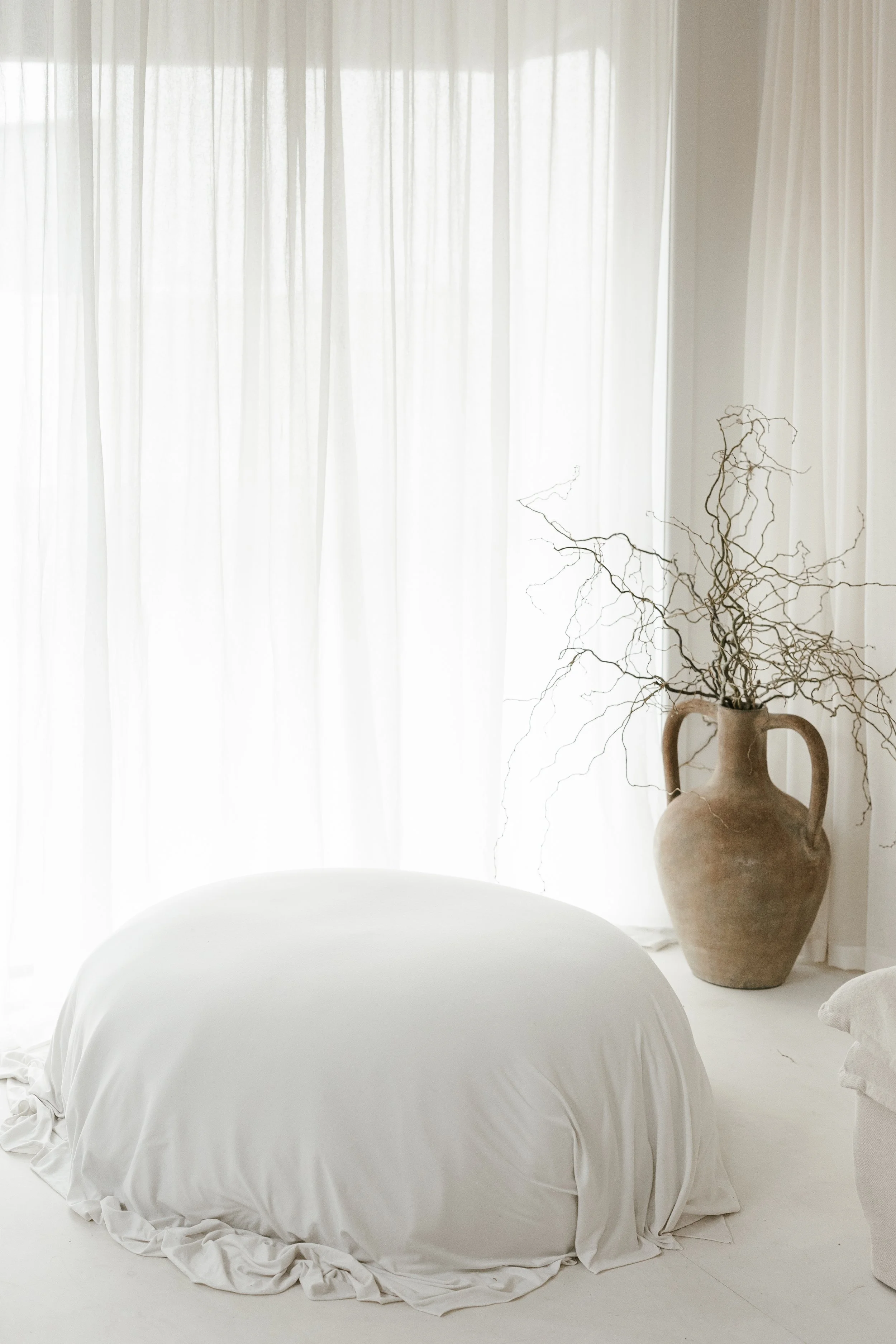 A bedroom with white curtains, a bed with white bedding, and a large beige vase with dried branches.