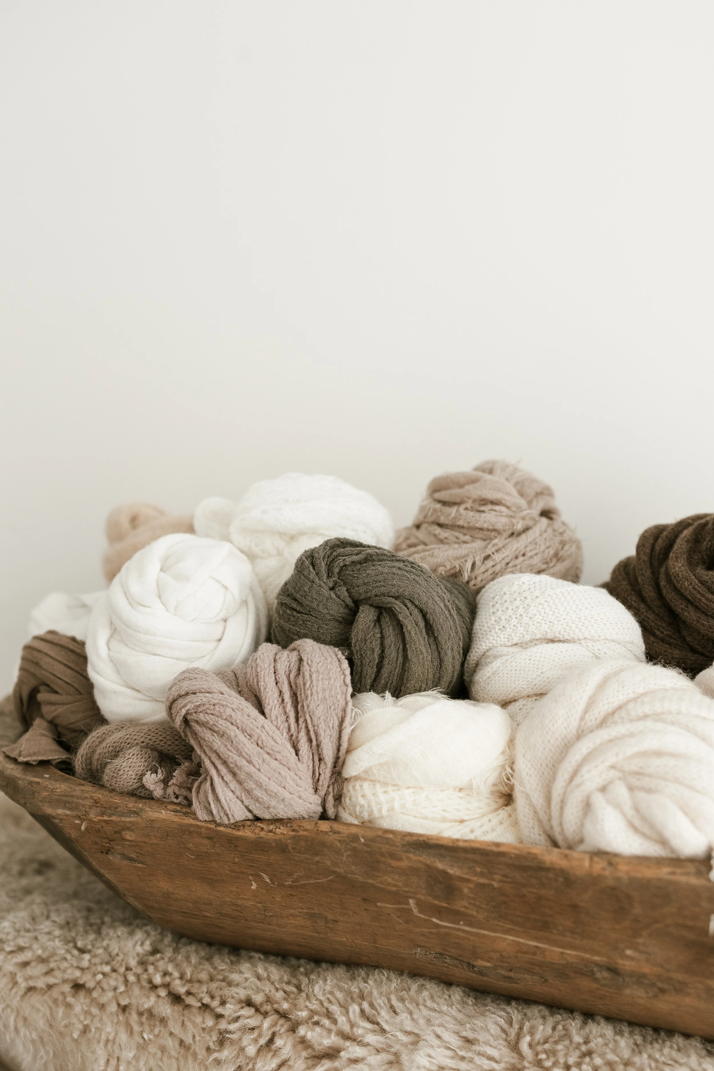 A wooden bowl filled with neatly rolled neutral-colored scarves and blankets, placed on a soft, fluffy beige carpet.