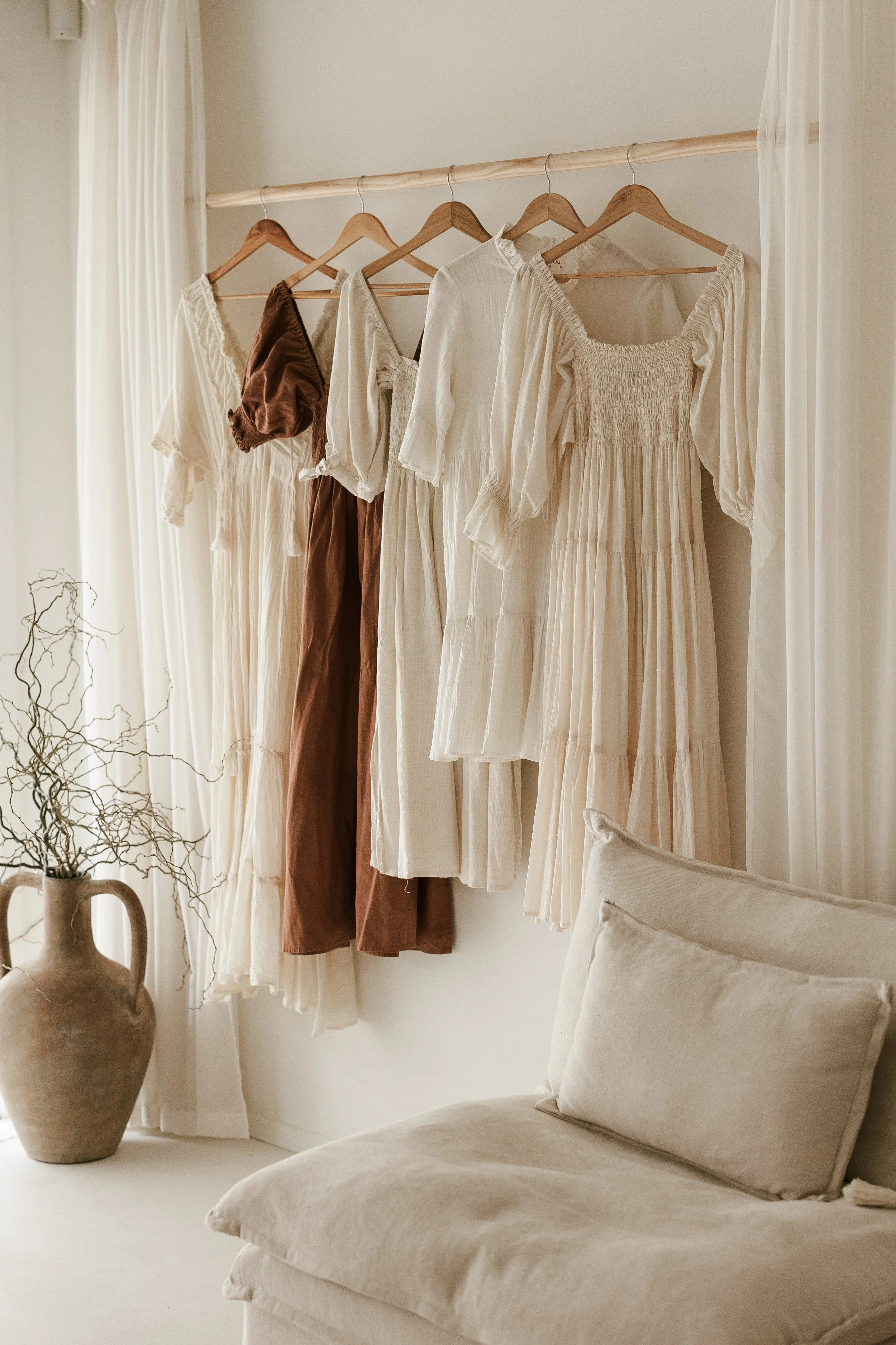 A row of hanging dresses and clothing on a wooden rod in a bright, neutral-colored room with natural light, a cream sofa in the foreground, and a large ceramic vase with dried branches on the left.