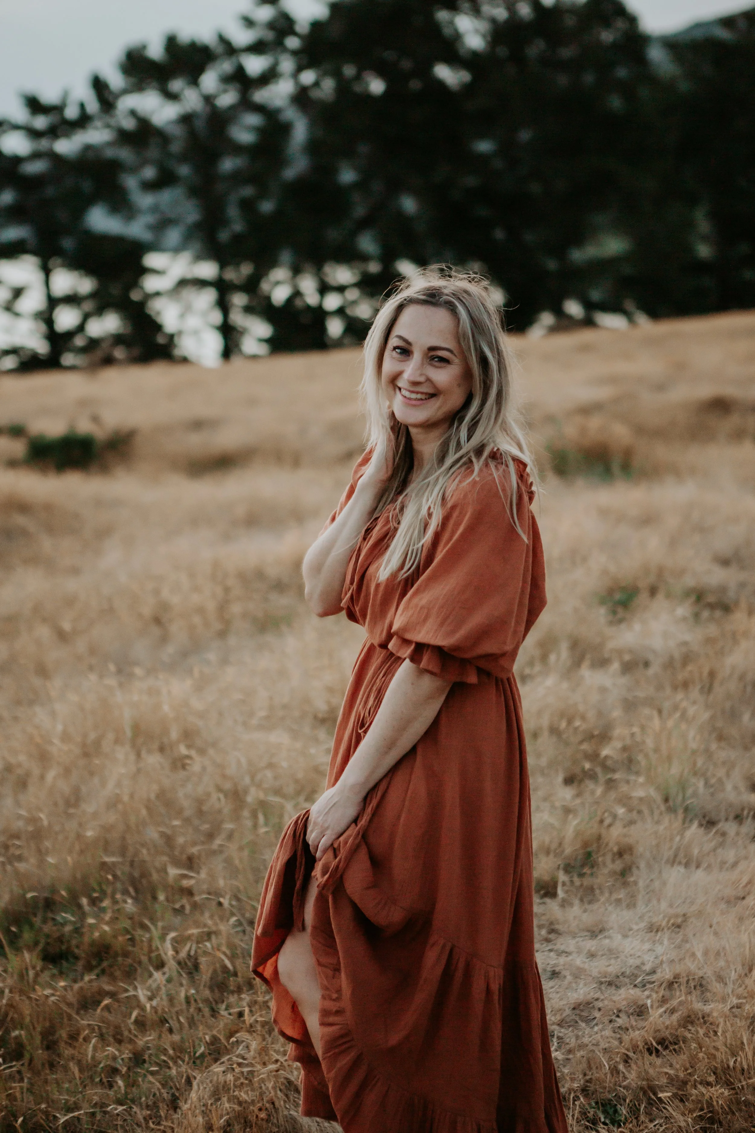 A woman with long blonde hair wearing a rust-colored dress stands in a grassy field, smiling and looking at the camera.