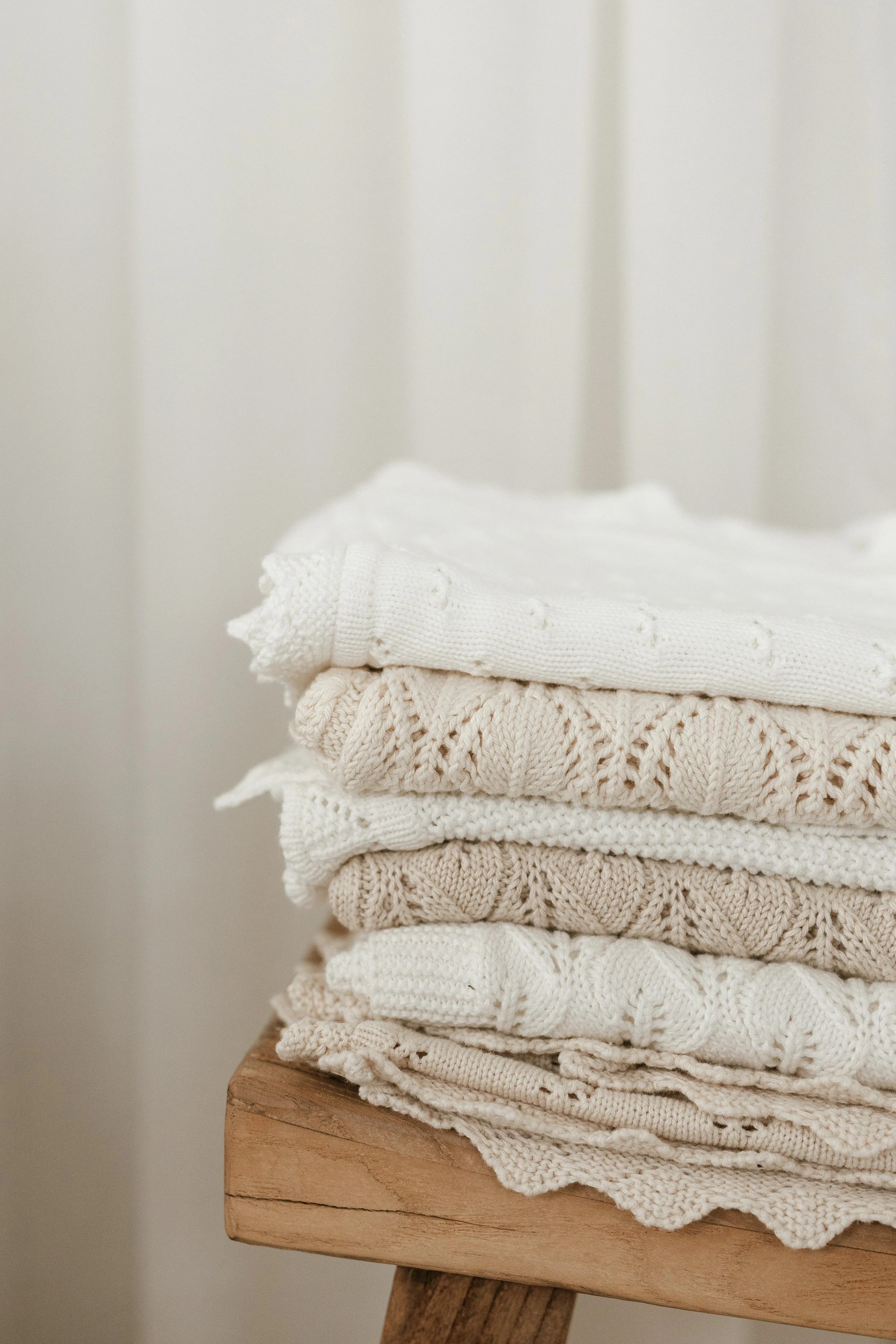 A stack of folded cream and white knitted sweaters on a wooden stool against a neutral background.