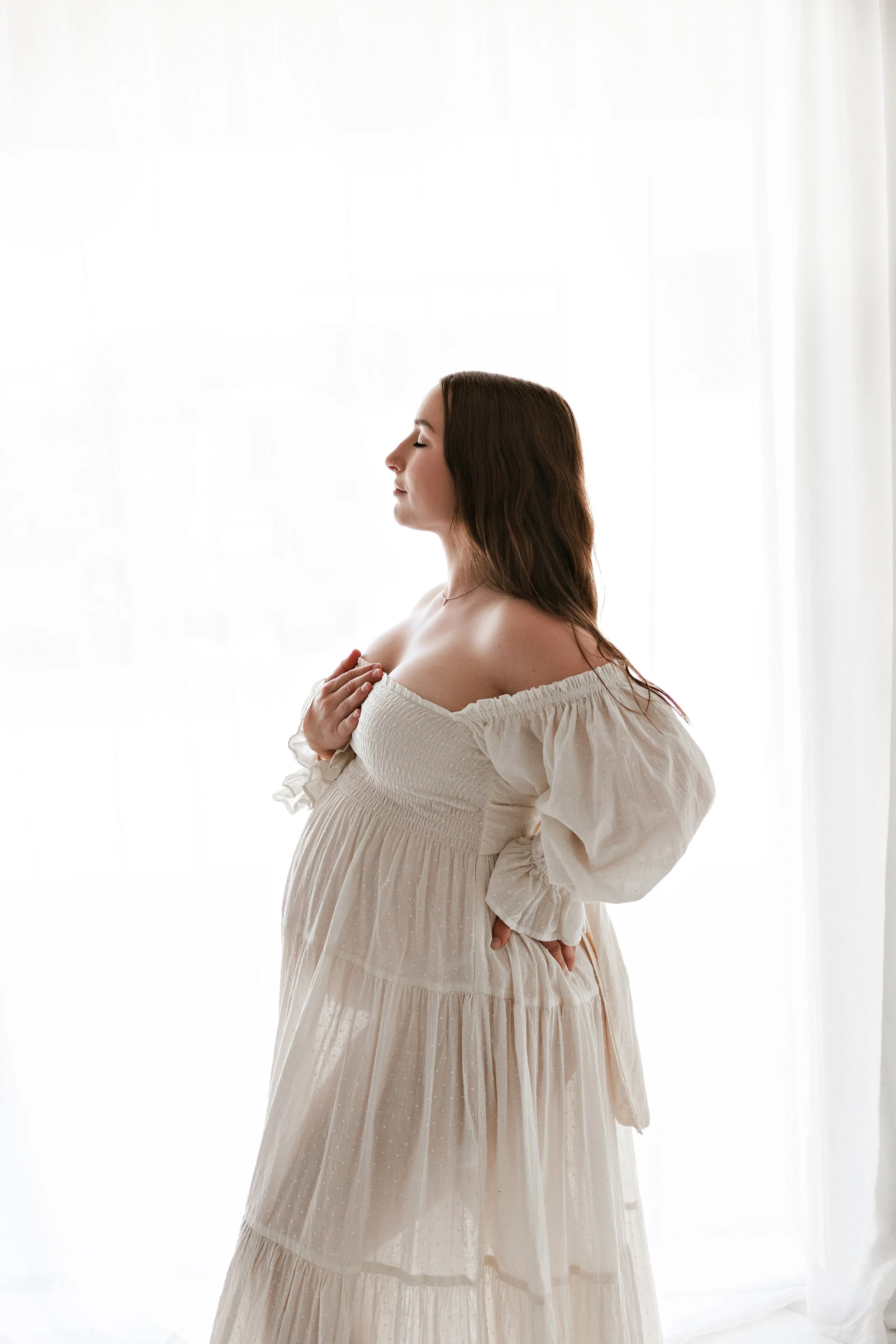 A pregnant woman with long brown hair, wearing a white off-the-shoulder dress, standing near a light-colored curtain with her eyes closed, gently holding her belly.