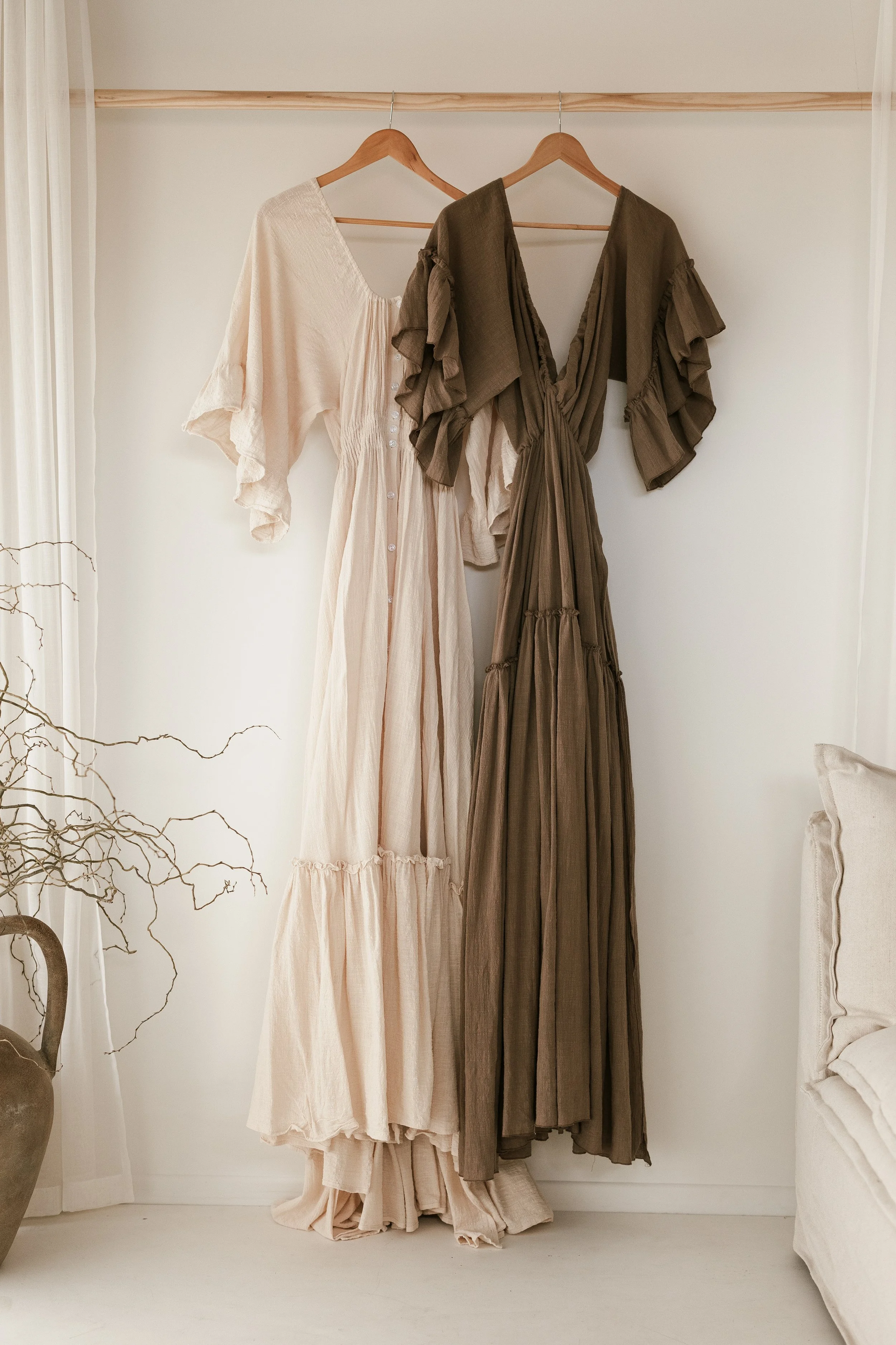 Two long dresses hanging on wooden hangers on a wooden rod against a white wall. One dress is cream-colored with buttons, and the other is brown with ruffled sleeves.