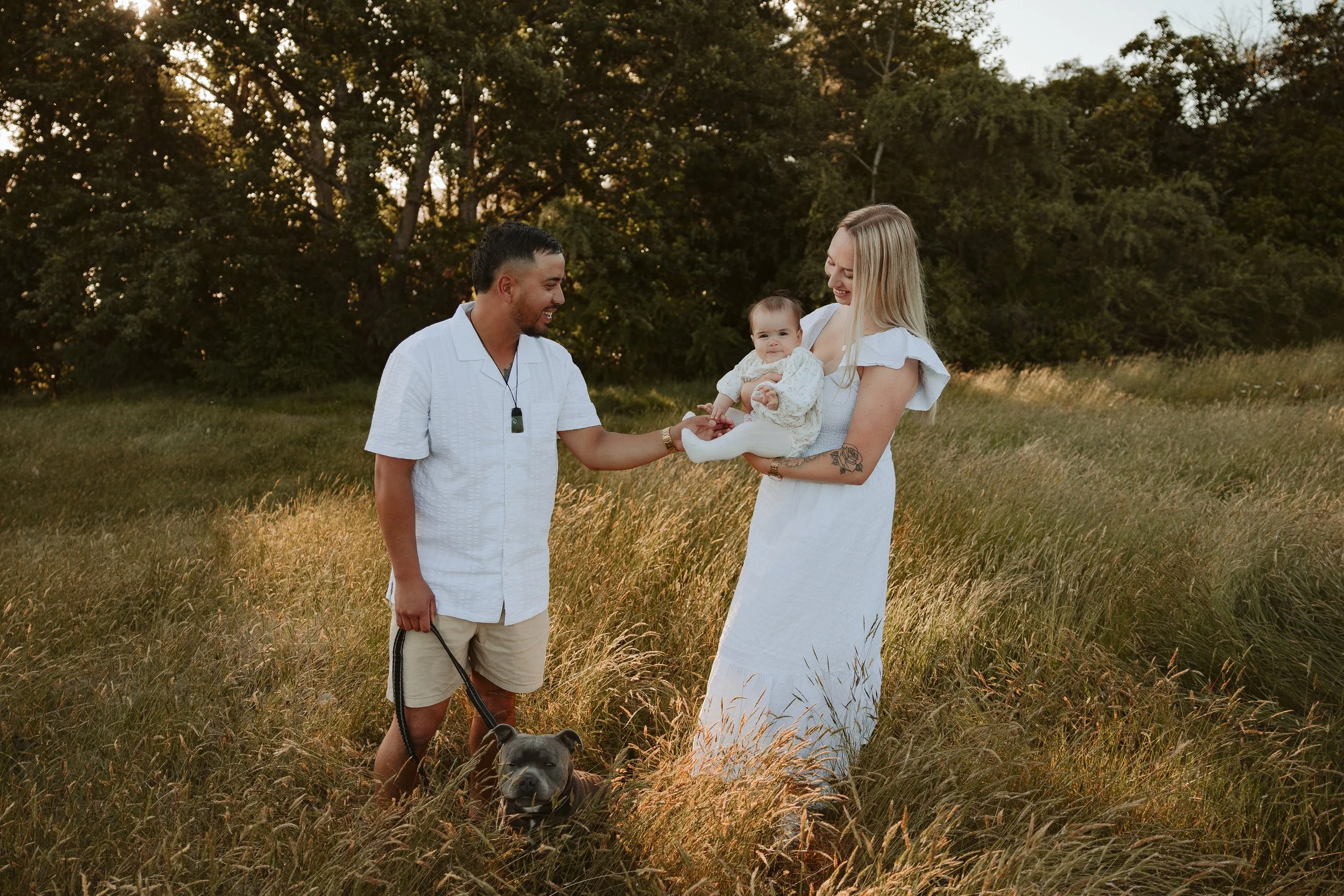 A family of three and their dog in a grassy field during sunset. The mother is holding their baby girl, while the father holds the dog's leash. They are all smiling and enjoying the moment.