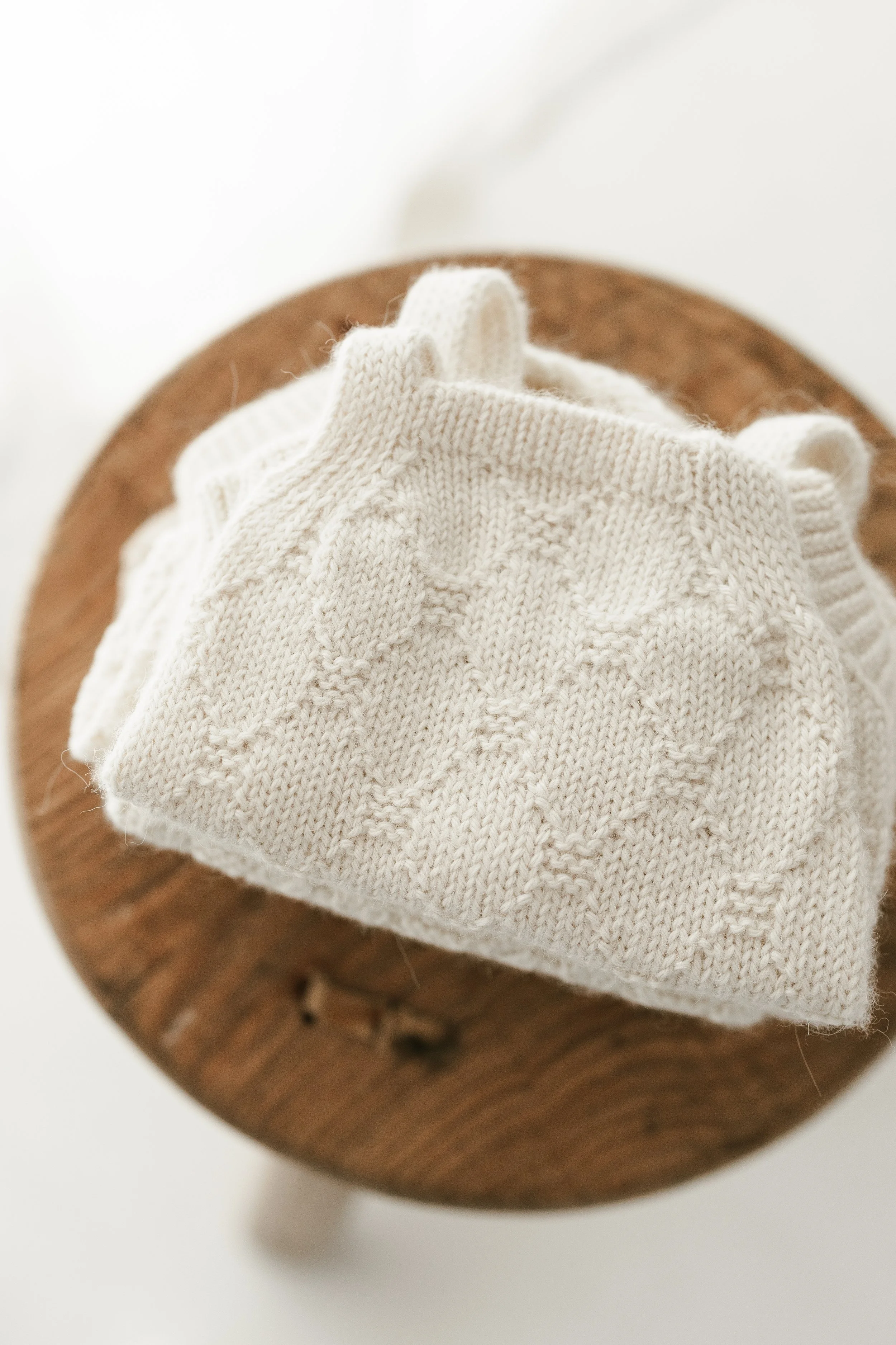 Close-up of a folded cream-colored cable-knit sweater with a leaf pattern, placed on a small, round wooden stool.