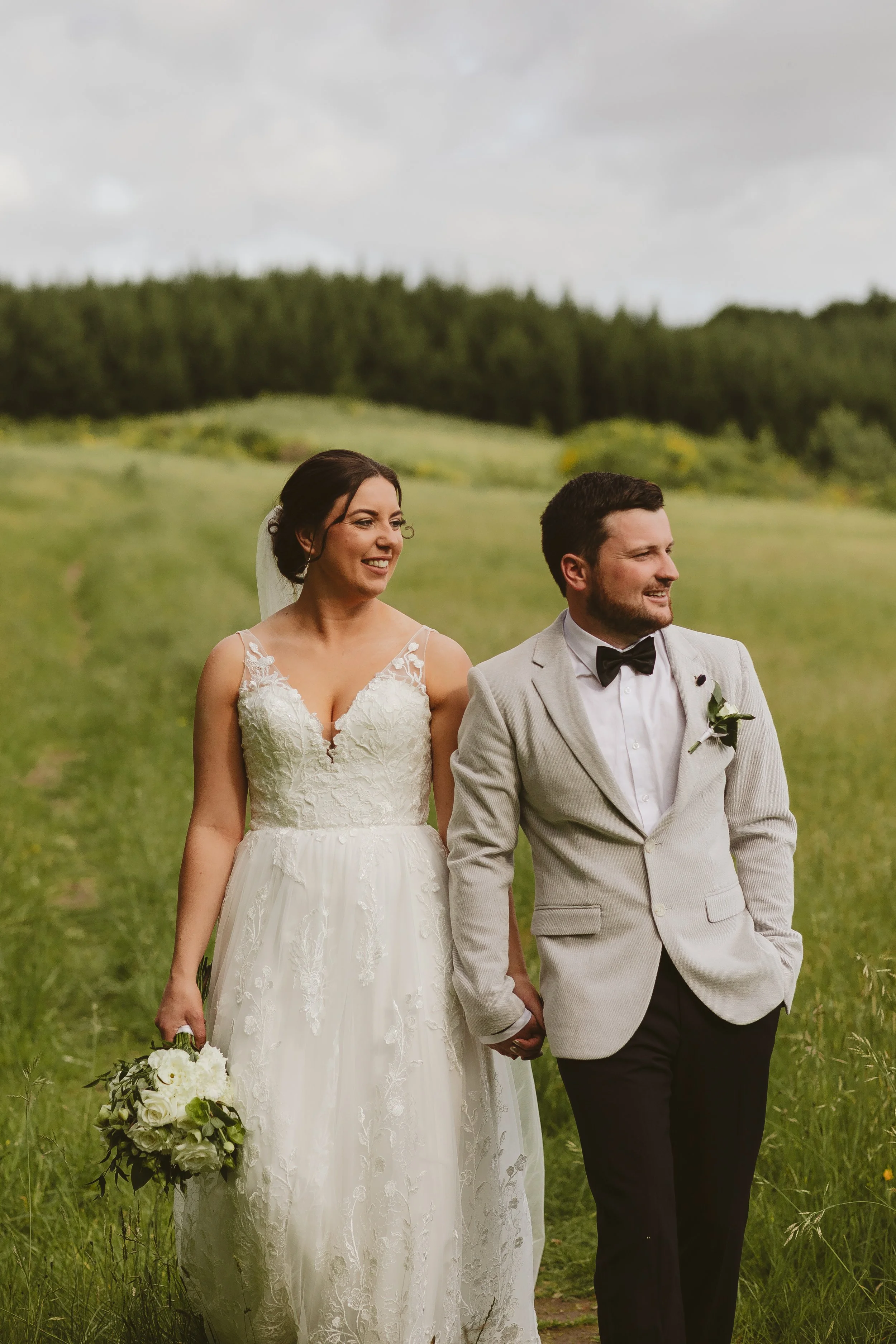 A bride and groom holding hands and walking in a green field with trees in the background on their wedding day.