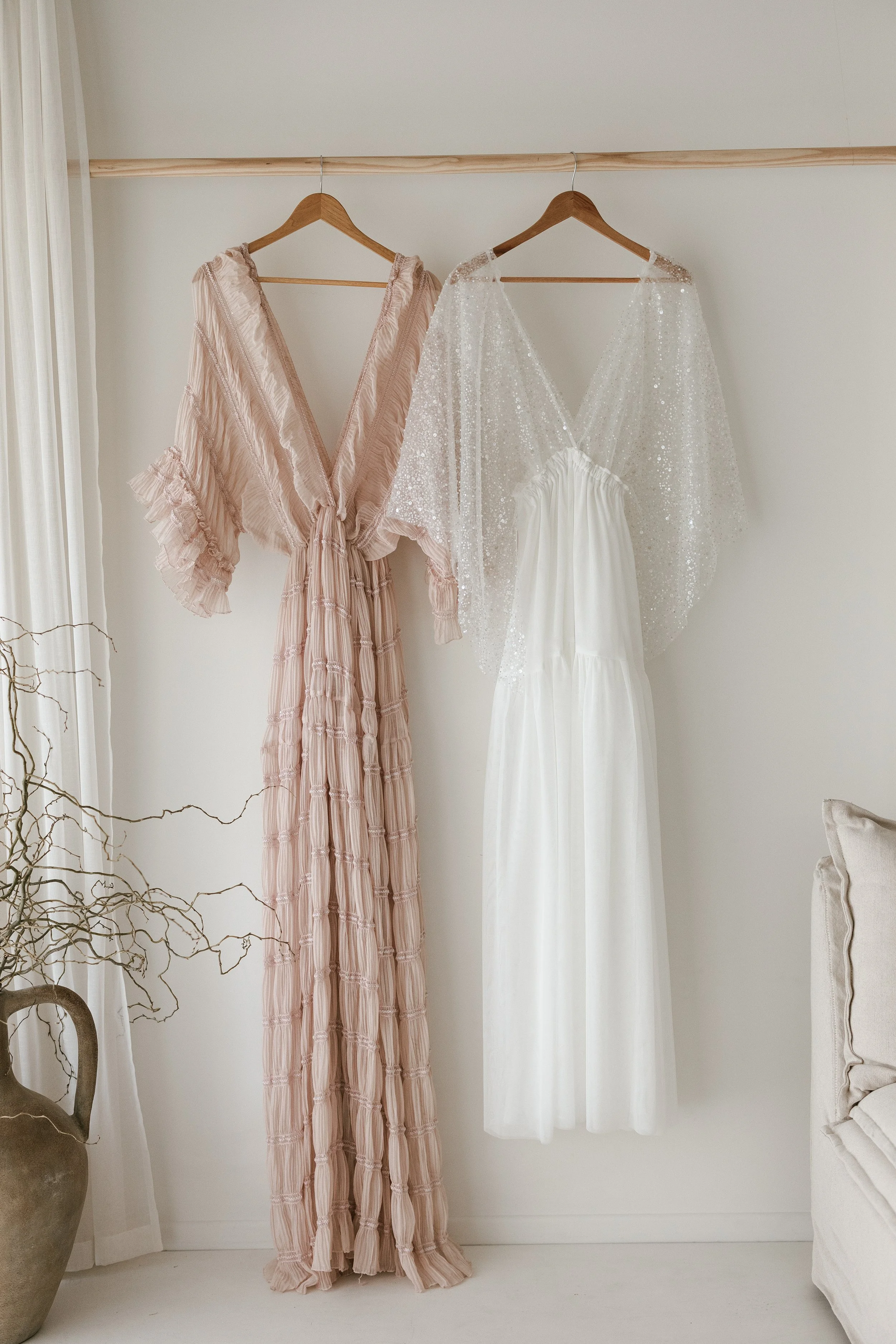 Two dresses hanging on a wooden rod; a pink, ruffled, tiered dress on the left, and a white, strapless, flowing dress with a sheer, sparkly cape on the right.