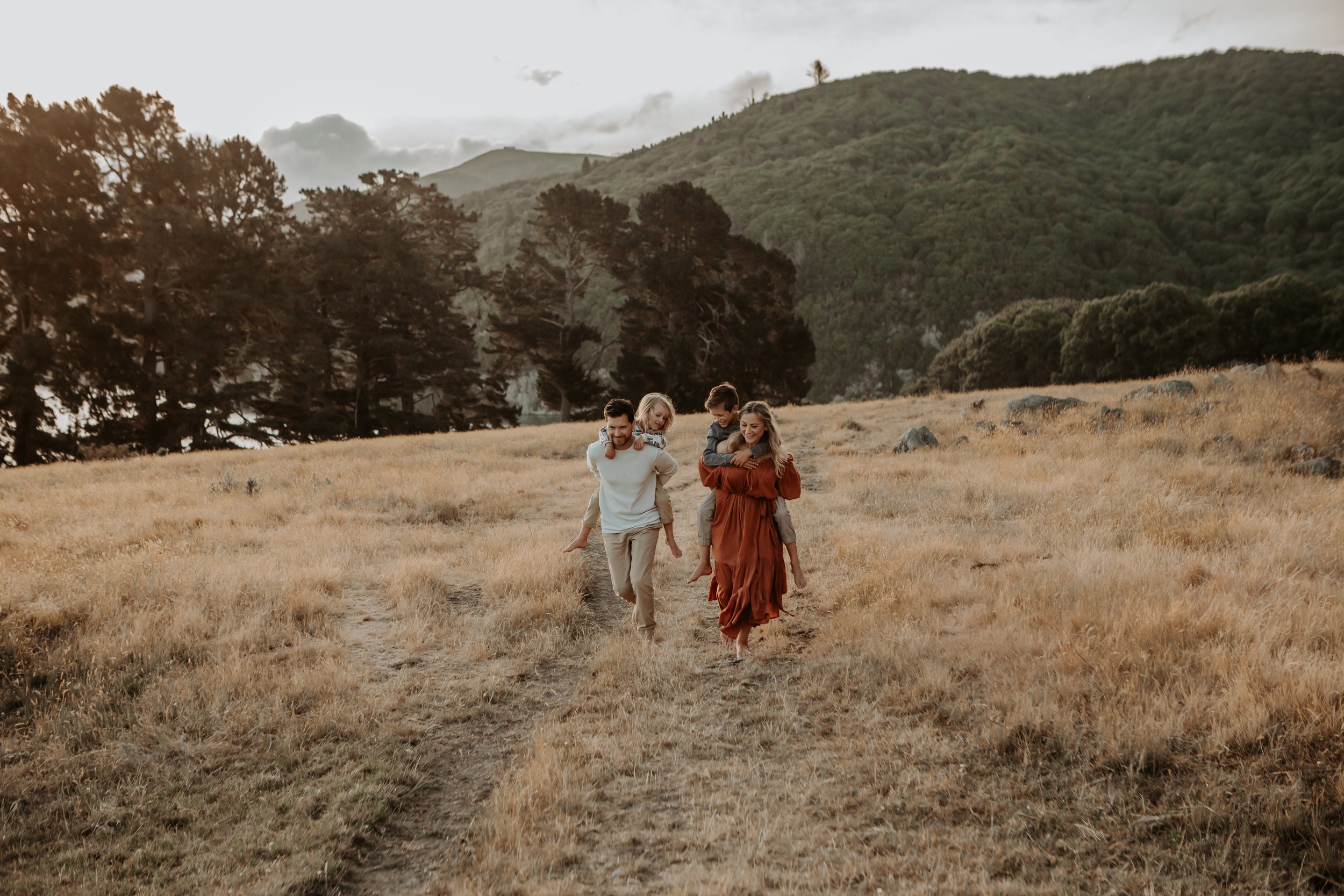 A family of four walking on a dirt trail through a grassy field with trees and mountains in the background during late afternoon or early evening.