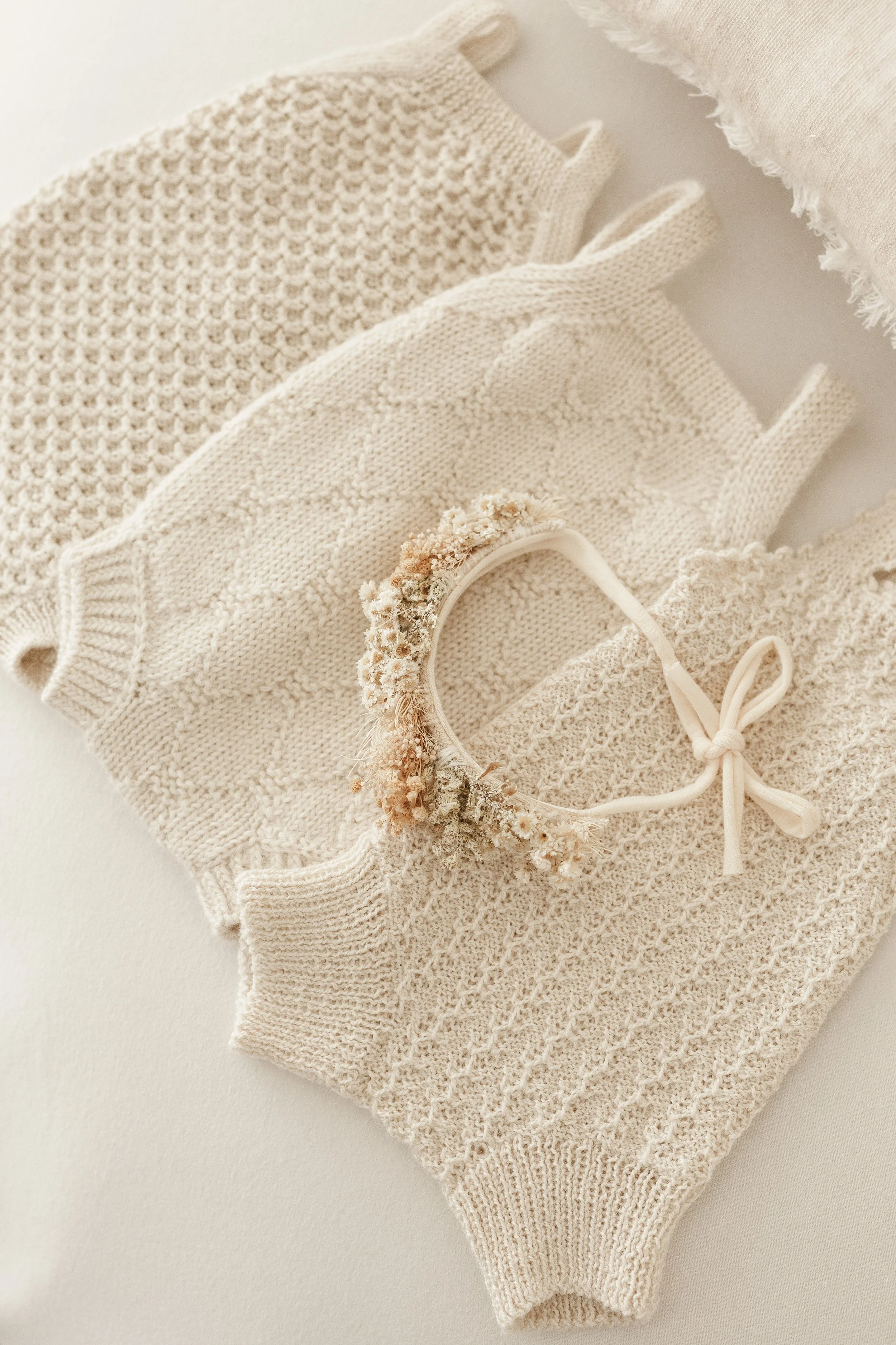 Three cream-colored knitted baby sweaters and a floral headband on a white surface.
