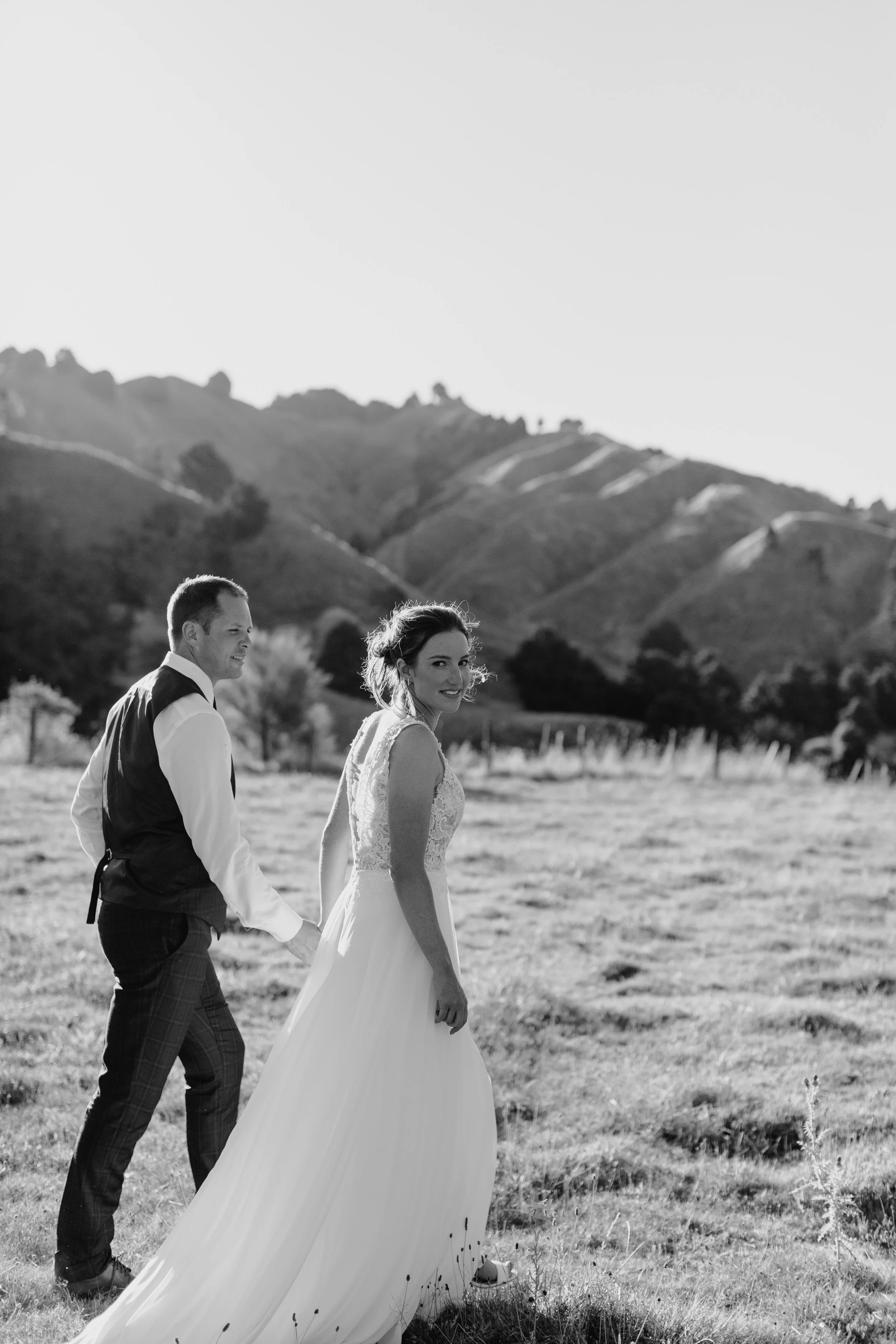 A black and white photograph of a couple in wedding attire walking outdoors in a grassy field with mountains in the background. The bride is wearing a white dress with lace and is smiling at the camera, while the groom is dressed in a vest and checkered pants, looking at the bride.