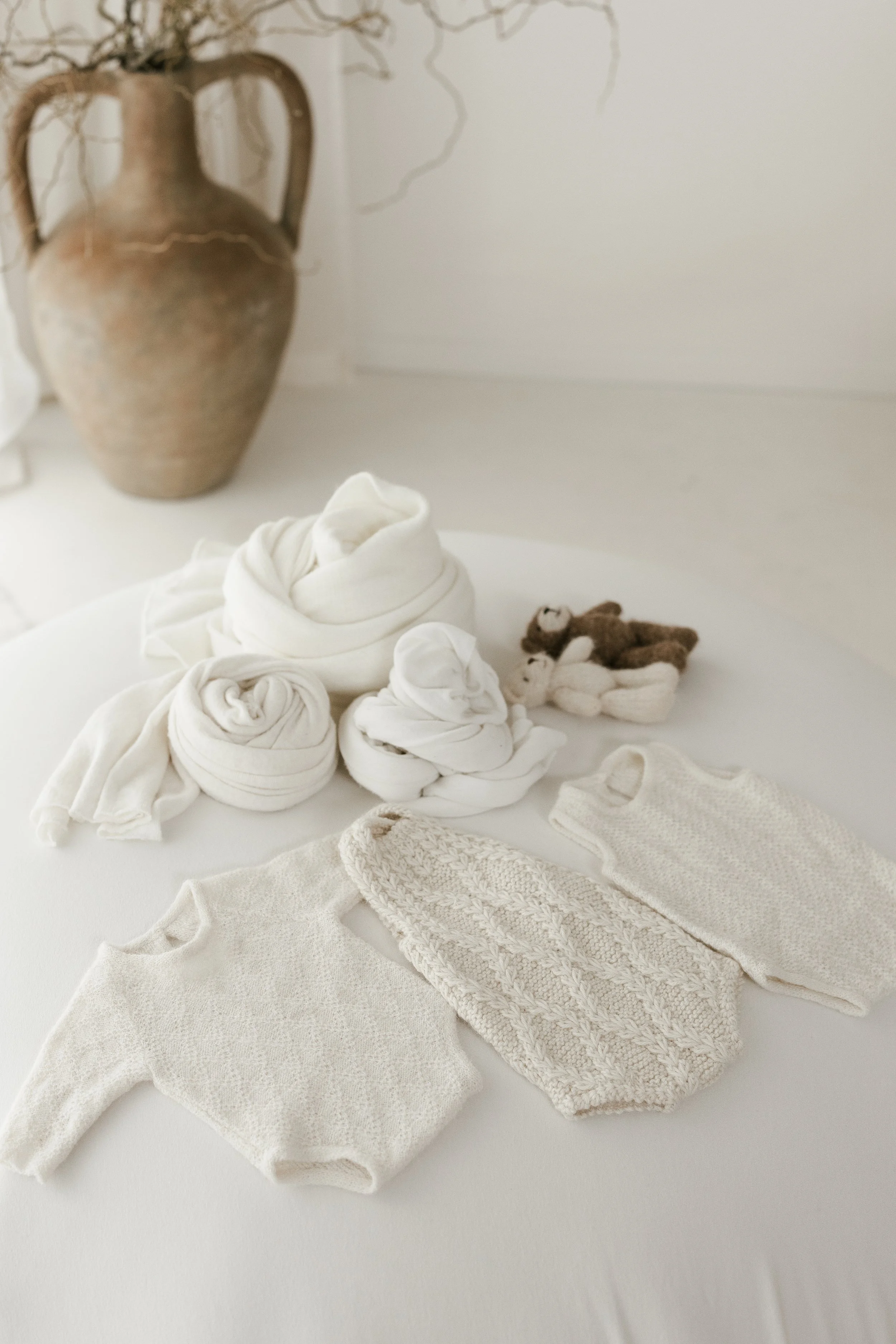 A collection of newborn clothing and accessories, including knitted and swaddled fabric bundles, small sweaters, and stuffed animals, all in white and beige tones, arranged on a white surface with a vintage vase in the background.