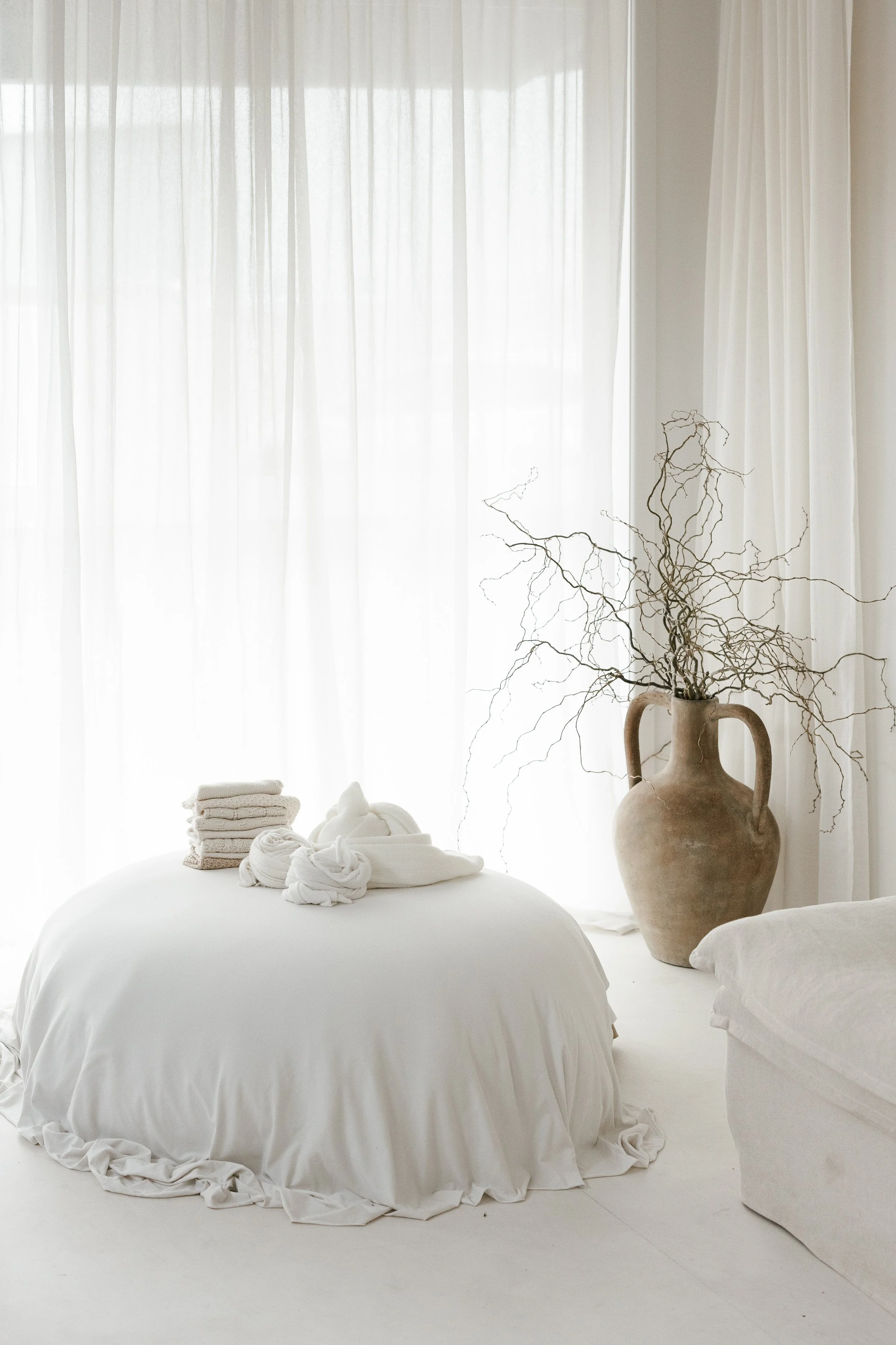 A bedroom with white curtains, a vase with bare branches, and a bed with neatly folded towels.