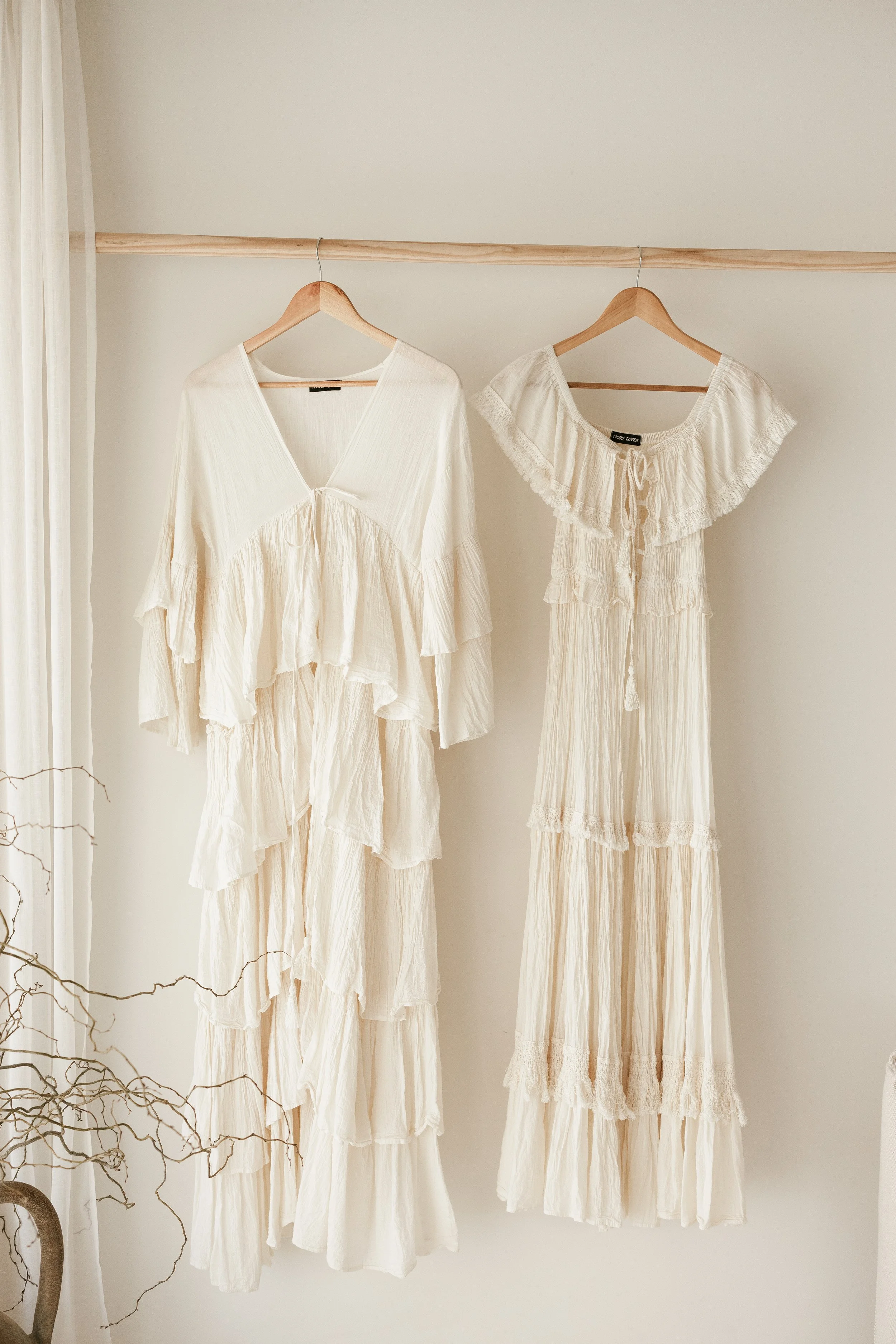 Two cream-colored, bohemian-style dresses hanging on wooden hangers on a wooden rod against a beige wall.
