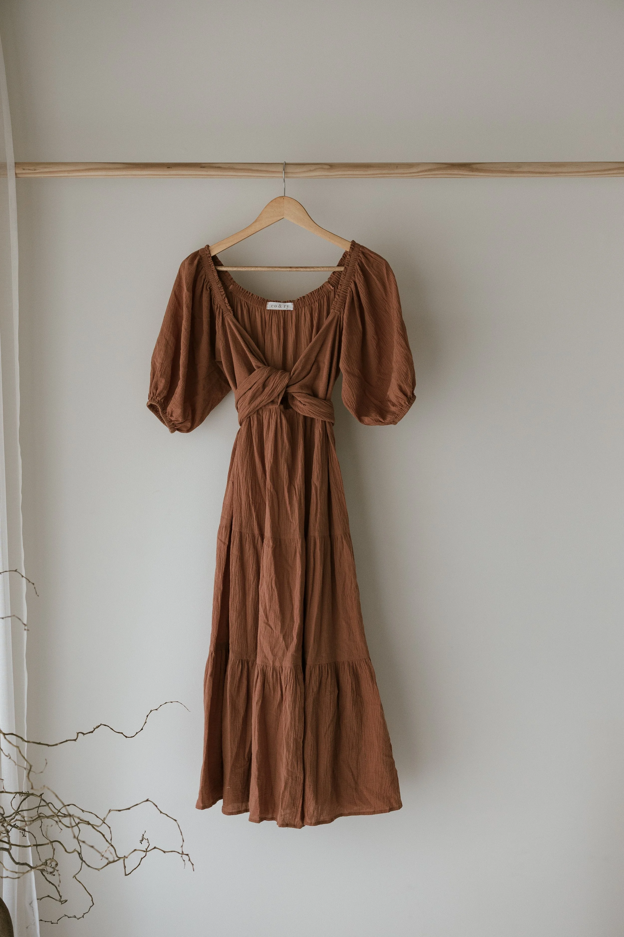 A brown dress with puffed sleeves hanging on a wooden hanger against a plain white wall.