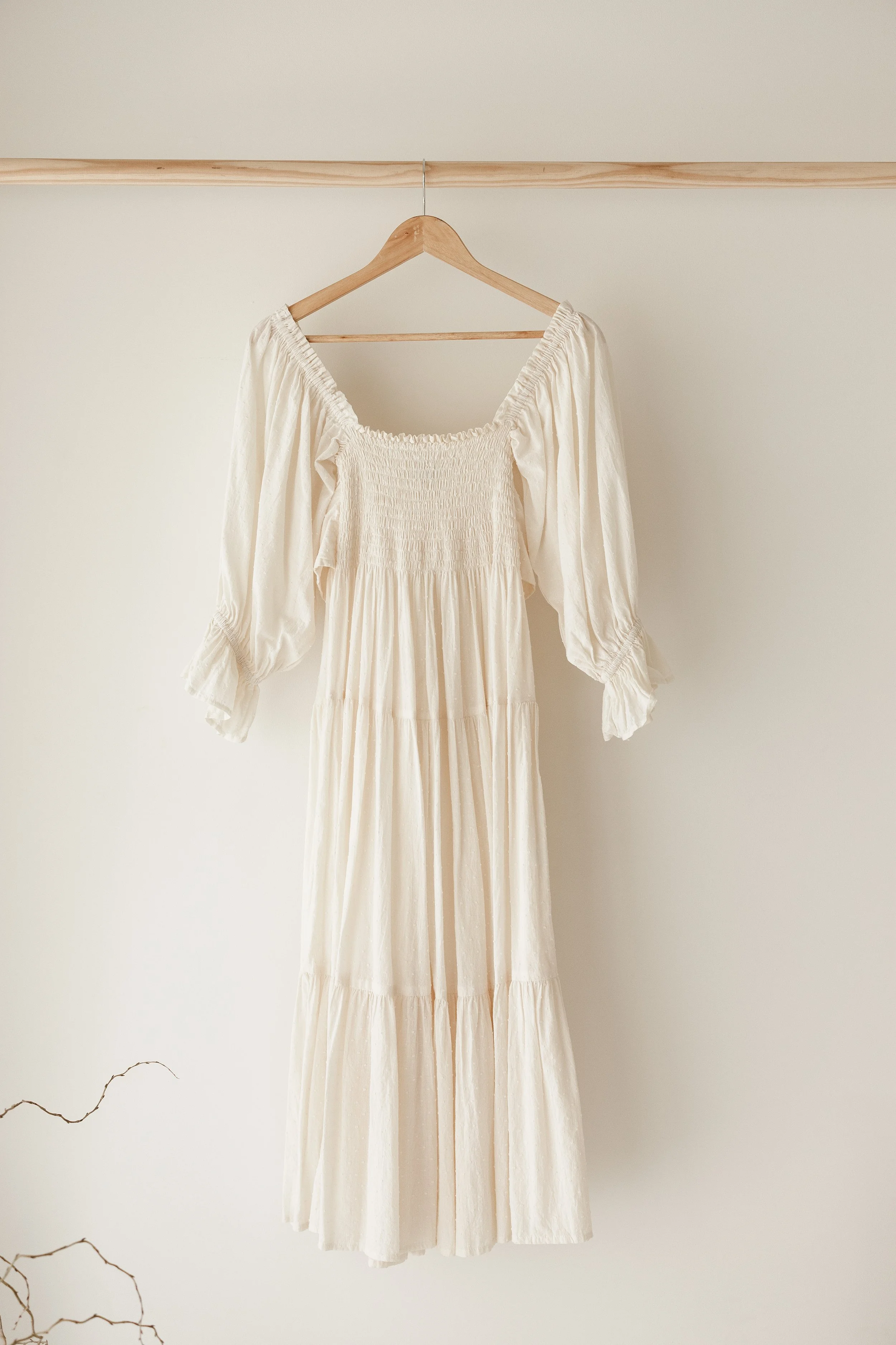 A long, off-white dress hanging on a wooden hanger against a plain wall.
