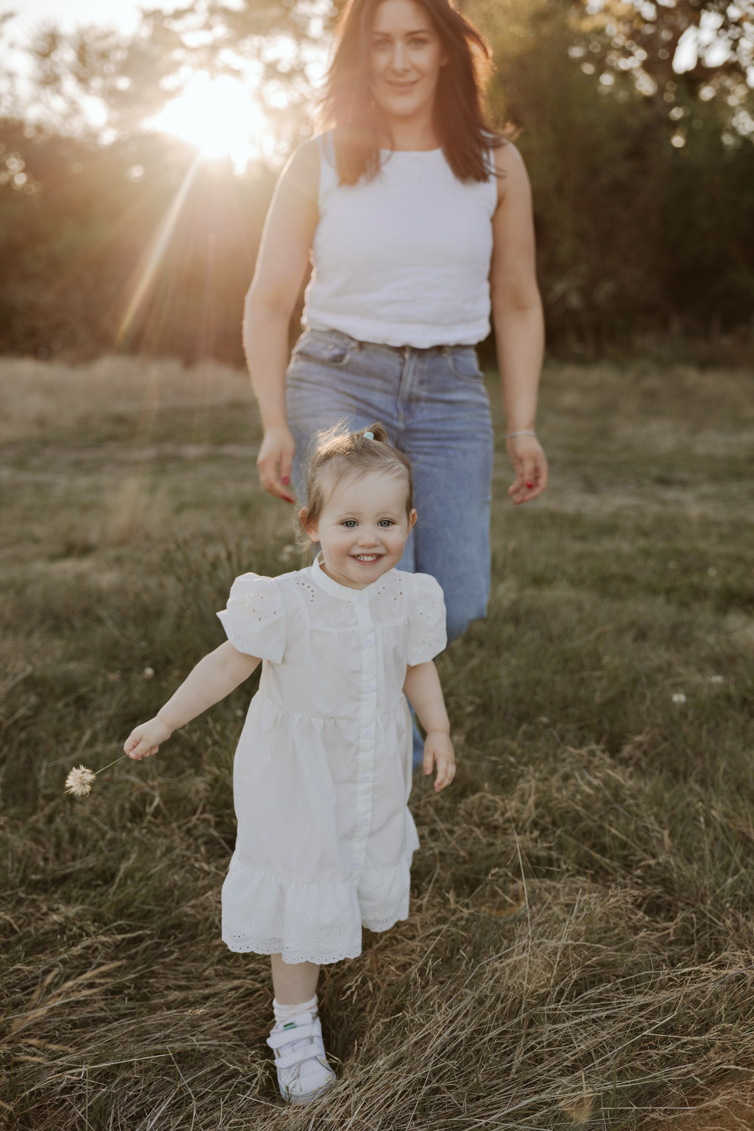 A woman and a young girl walking outdoors in a field at sunset. The woman is smiling and wearing a white sleeveless top and blue jeans. The young girl is smiling, wearing a white dress, white socks, and sneakers, and holding a small flower dandelion in her right hand.